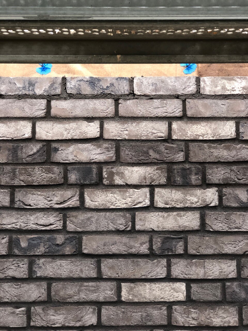 Grau-braune Backsteinmauer mit einer oberen Metallleiste; zwei blaue Kunststoffteile am Rand.