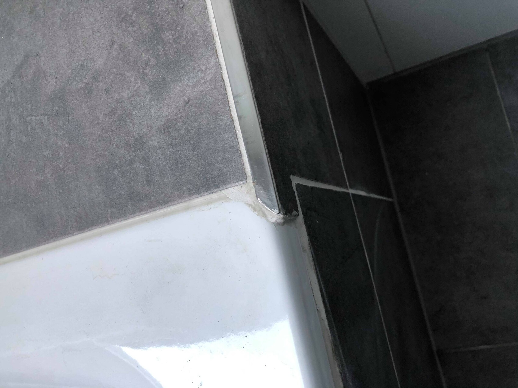 Eckkante der Badewanne: silberner Rand zwischen dunklen Fliesen und weißer Badewannenoberfläche.