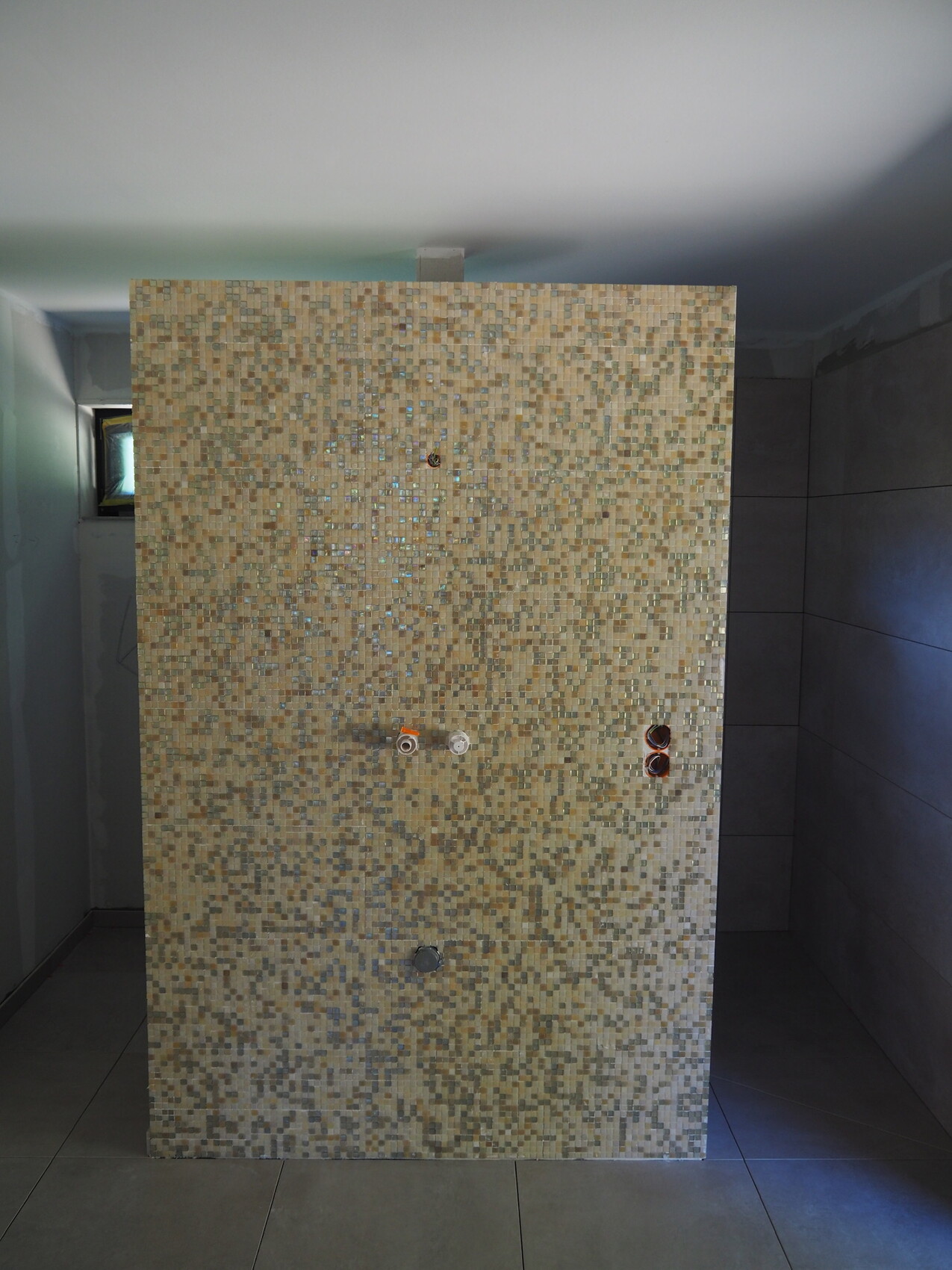 Beige Mosaikfliesenwand aus kleinen Fliesen im Badezimmer, Wasseranschlüsse sichtbar.