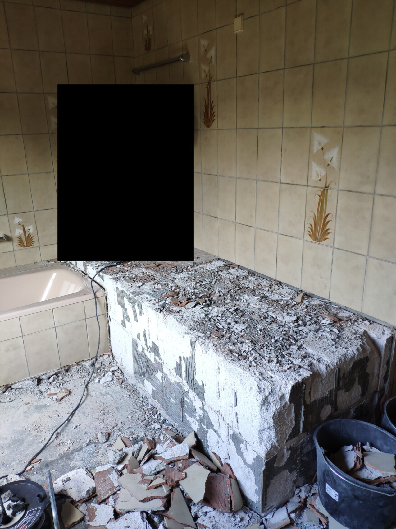Badezimmer im Abriss: großer Betonblock, bröckelnder Wandputz, Fliesenbruch und Schutt auf dem Boden.