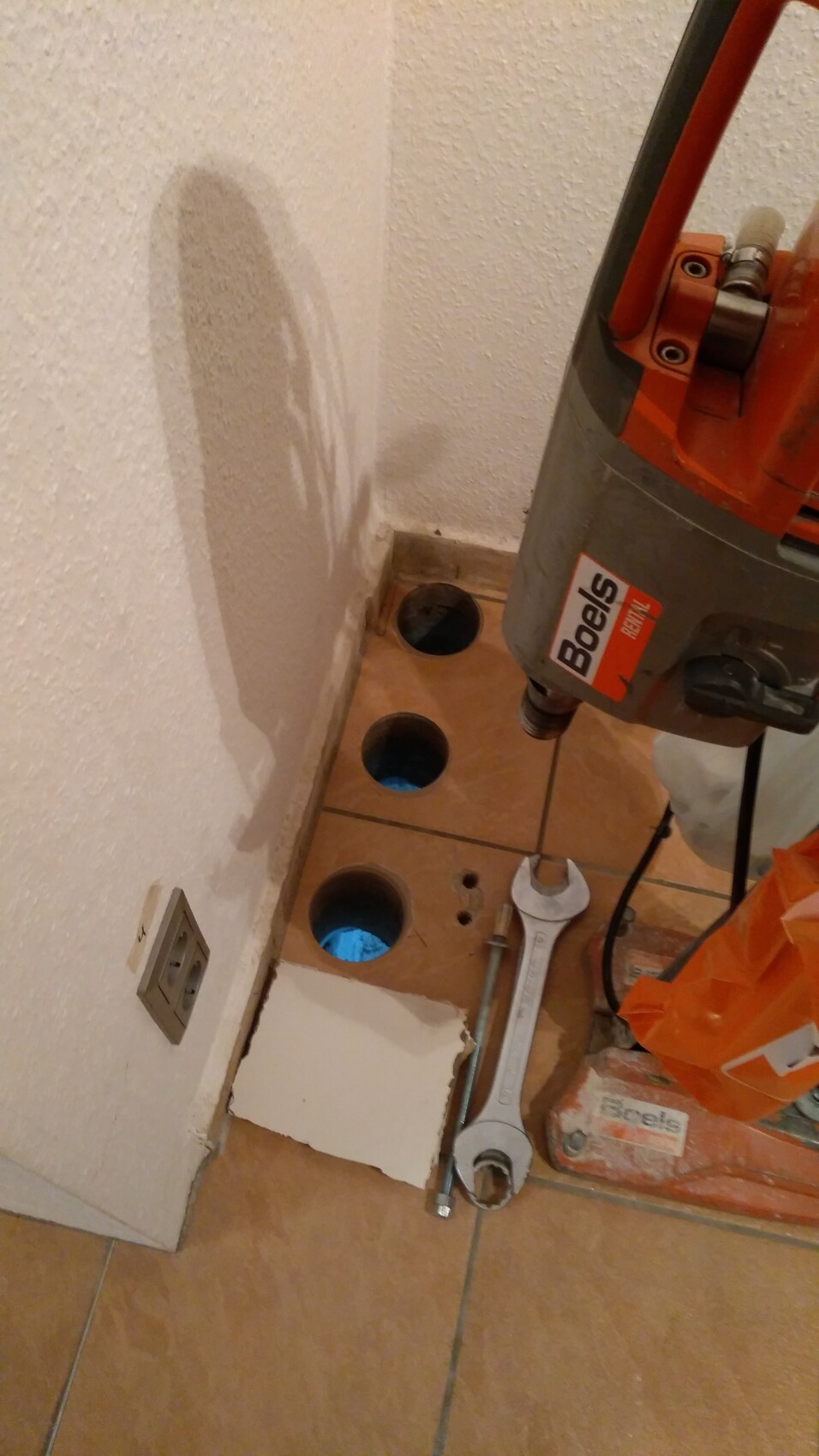 Bohrhammer neben drei runden Löchern in braunen Fliesen; Werkzeuge liegen bereit.
