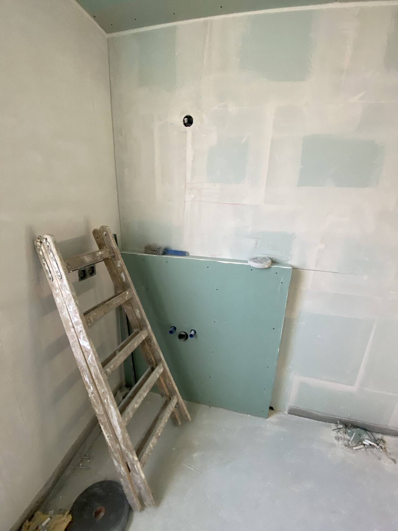 Unfertiges Badezimmer: grüne Installationsplatte, Leiter davor, Wasseranschlüsse sichtbar, Staub.