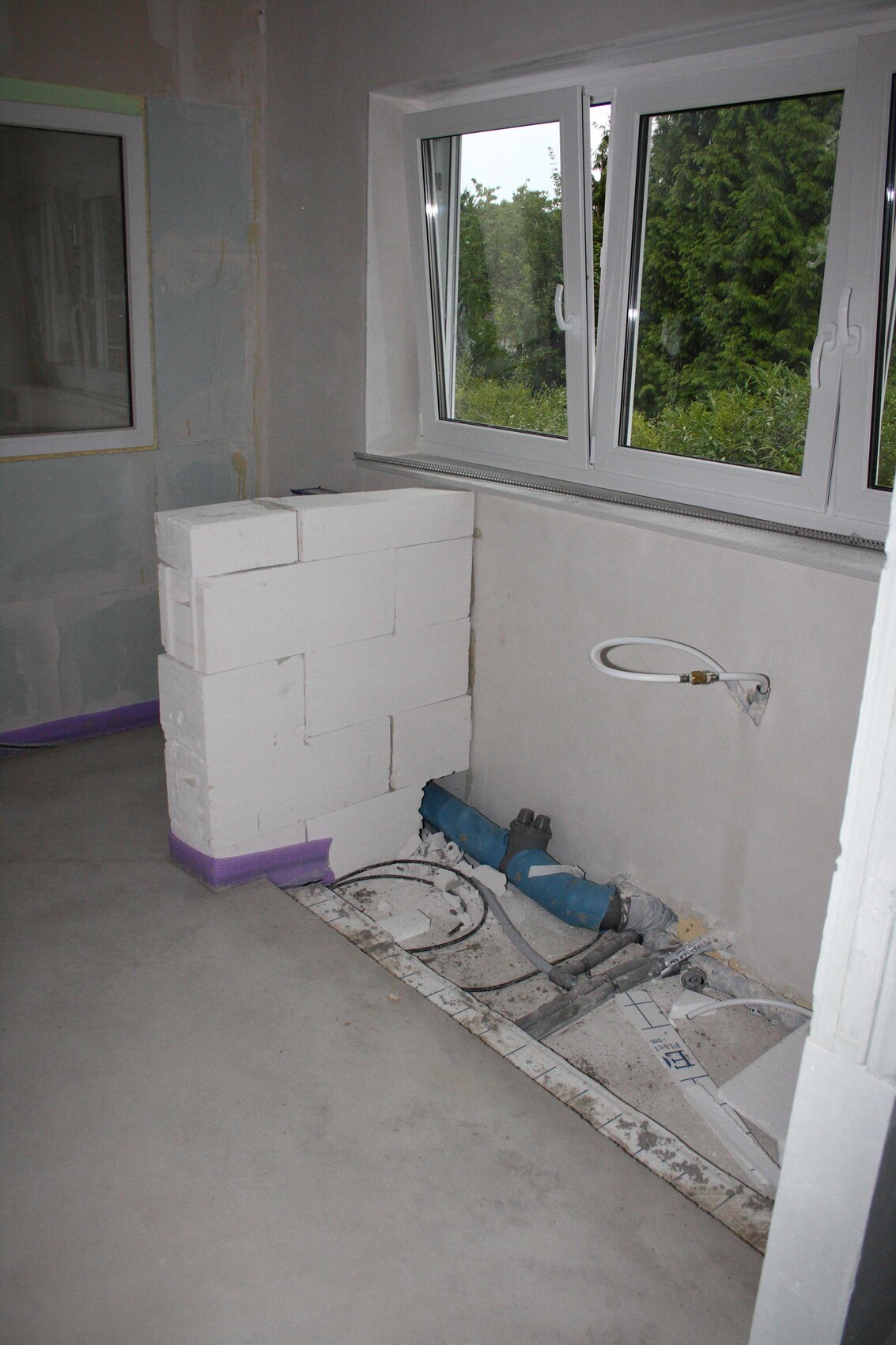 Innentrennwand aus Styroporblöcken neben Fenster; Rohre und Kabel auf dem Boden sichtbar.