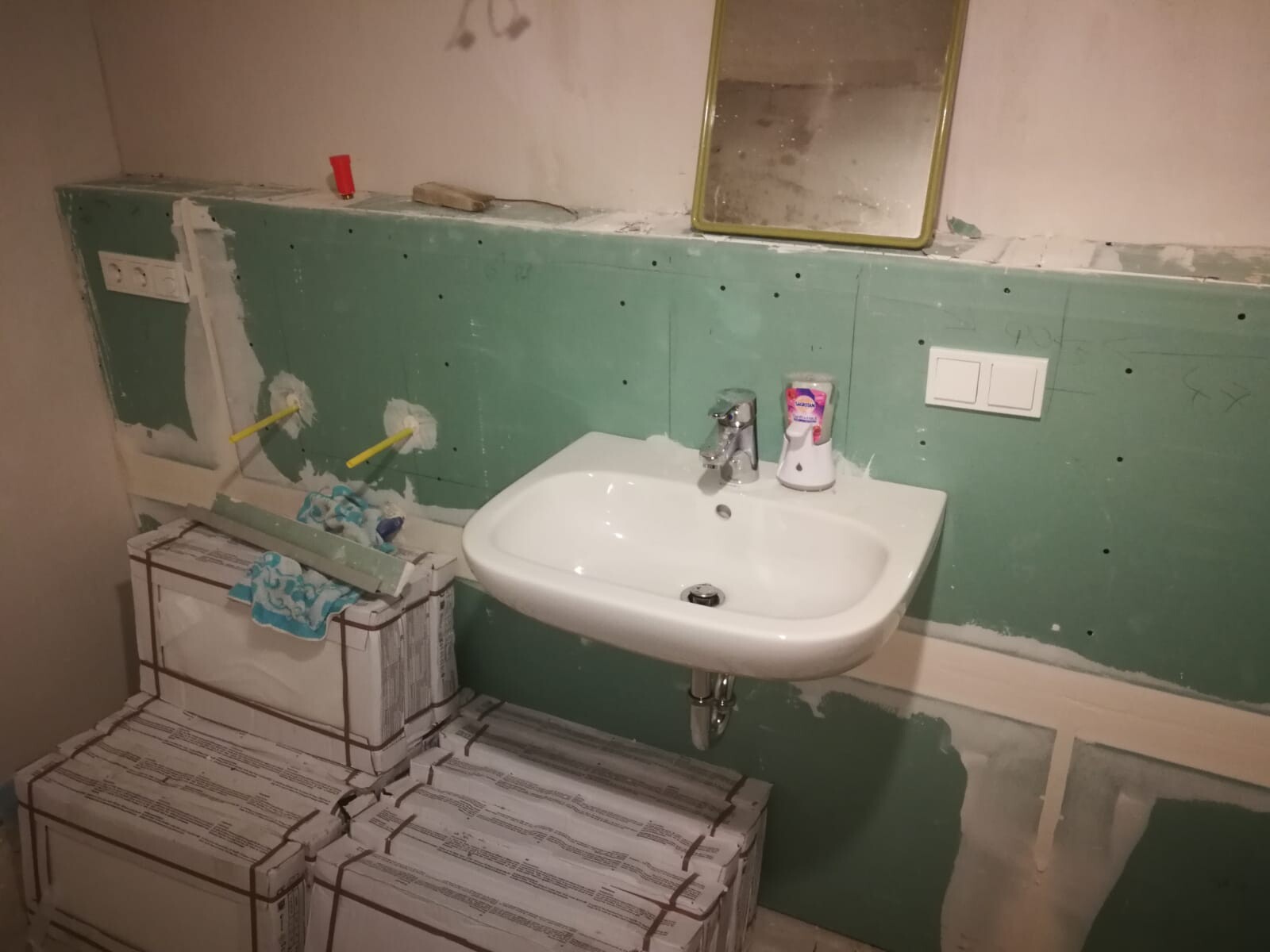 Unter Renovierung: Weißes Waschbecken an grüner Wand, gelb gerahmter Spiegel, Baupakete auf Boden.