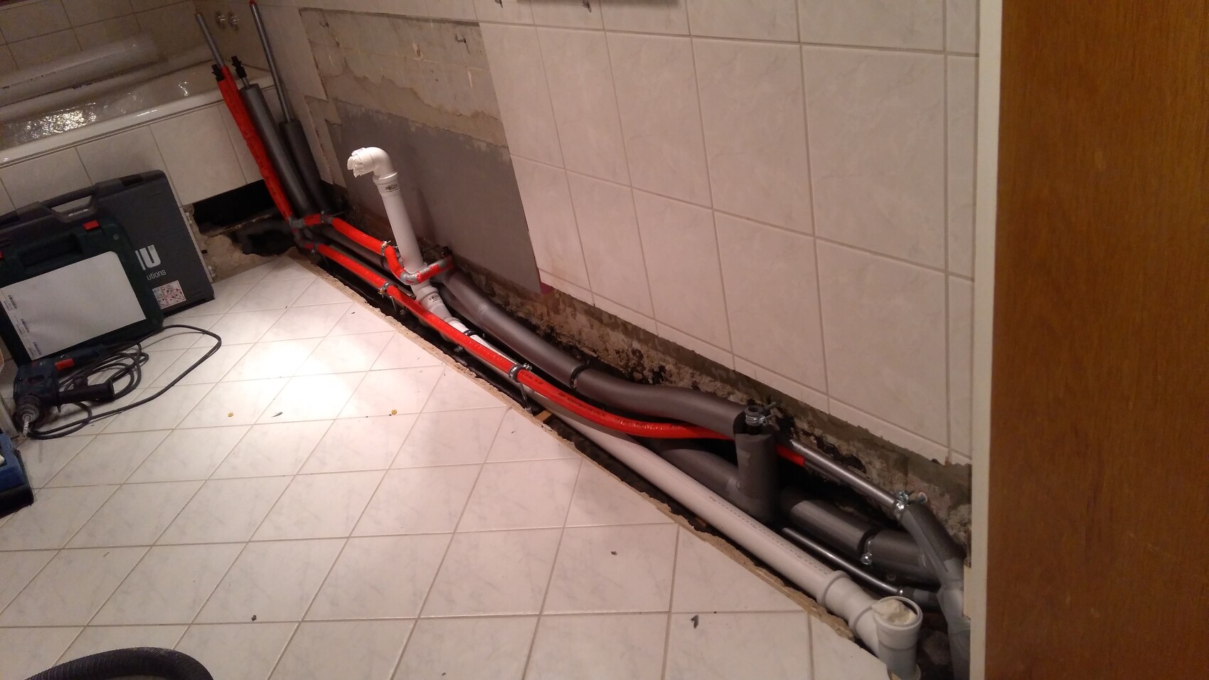 Unteres Badezimmer: rote PEX-Rohre, graue Abflussrohre, freigelegte Bodenöffnung und Werkzeugkasten.