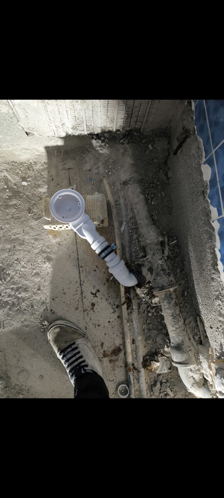 Baustellenboden mit weißem PVC-Abflussrohr, Schellen und Betonresten; Schuh im Vordergrund.