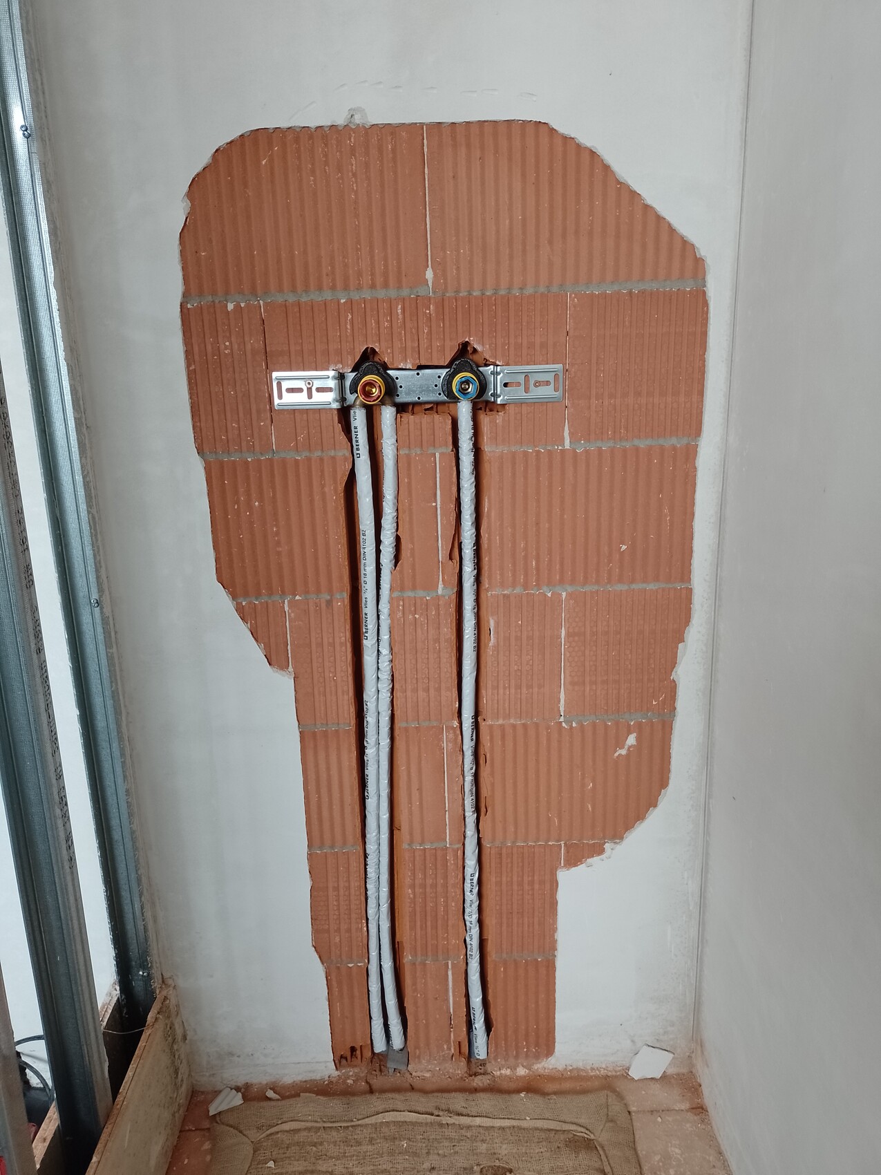 Freigelegte rote Ziegelwand mit zwei isolierten Wasserleitungen (Warm/Kalt) an Halterung.