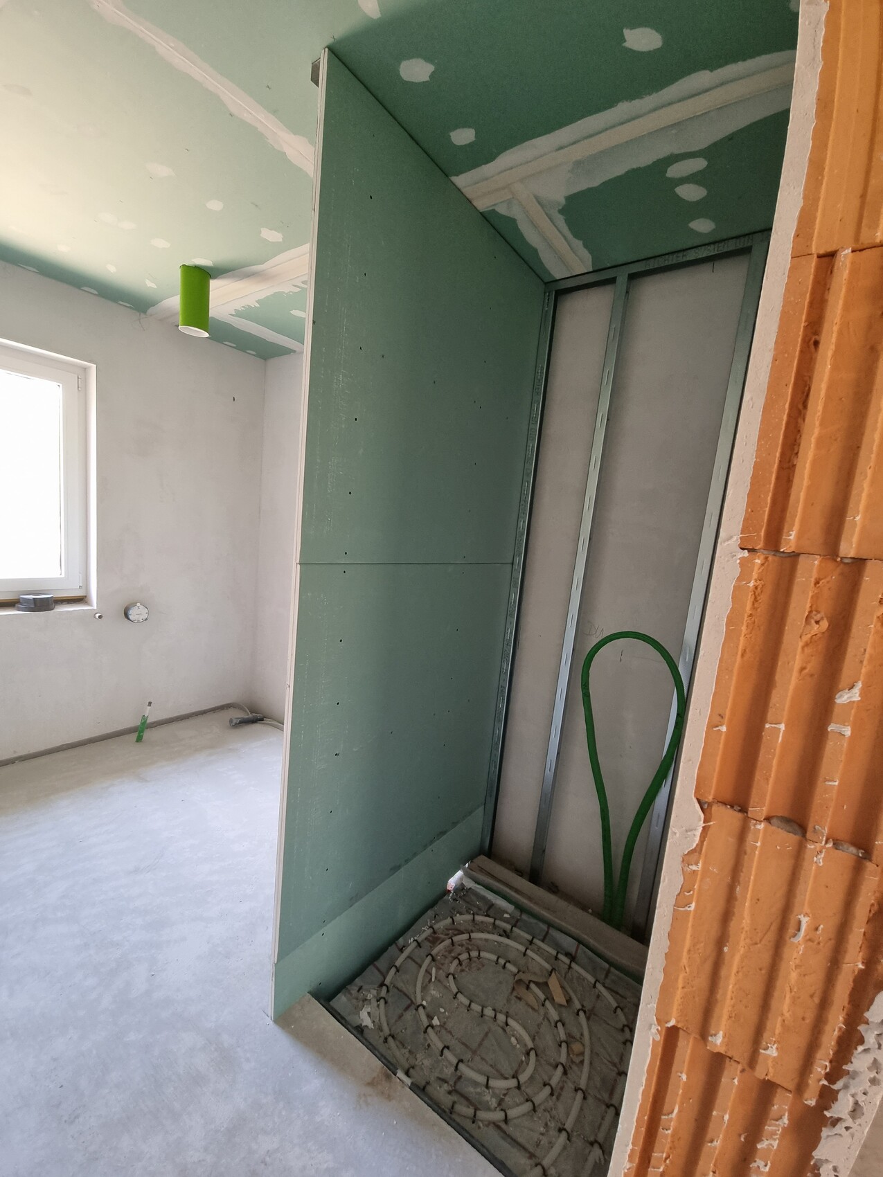 Rohbauzimmer mit grünem Trockenbau, Fenster links, grüne Rohre, Bodenöffnung.