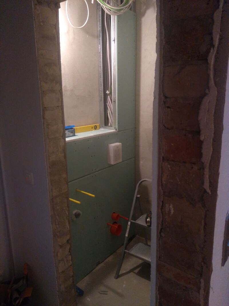Unfertiger Installationsraum mit grünem Trockenbau, Leiter, sichtbaren Kabeln und Backsteinwand.