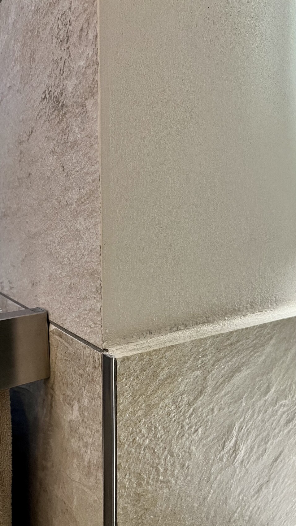 Eckbereich einer Dusche: linke Fliesen, rechte glatte Wand, unten Steinboden, Metallrahmen Glaswand.