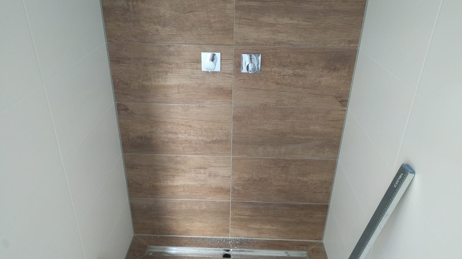 Badezimmerwand mit braunen Holzoptikfliesen, zwei silberne Schalter, Bodeneinbau-Duschschiene sichtbar