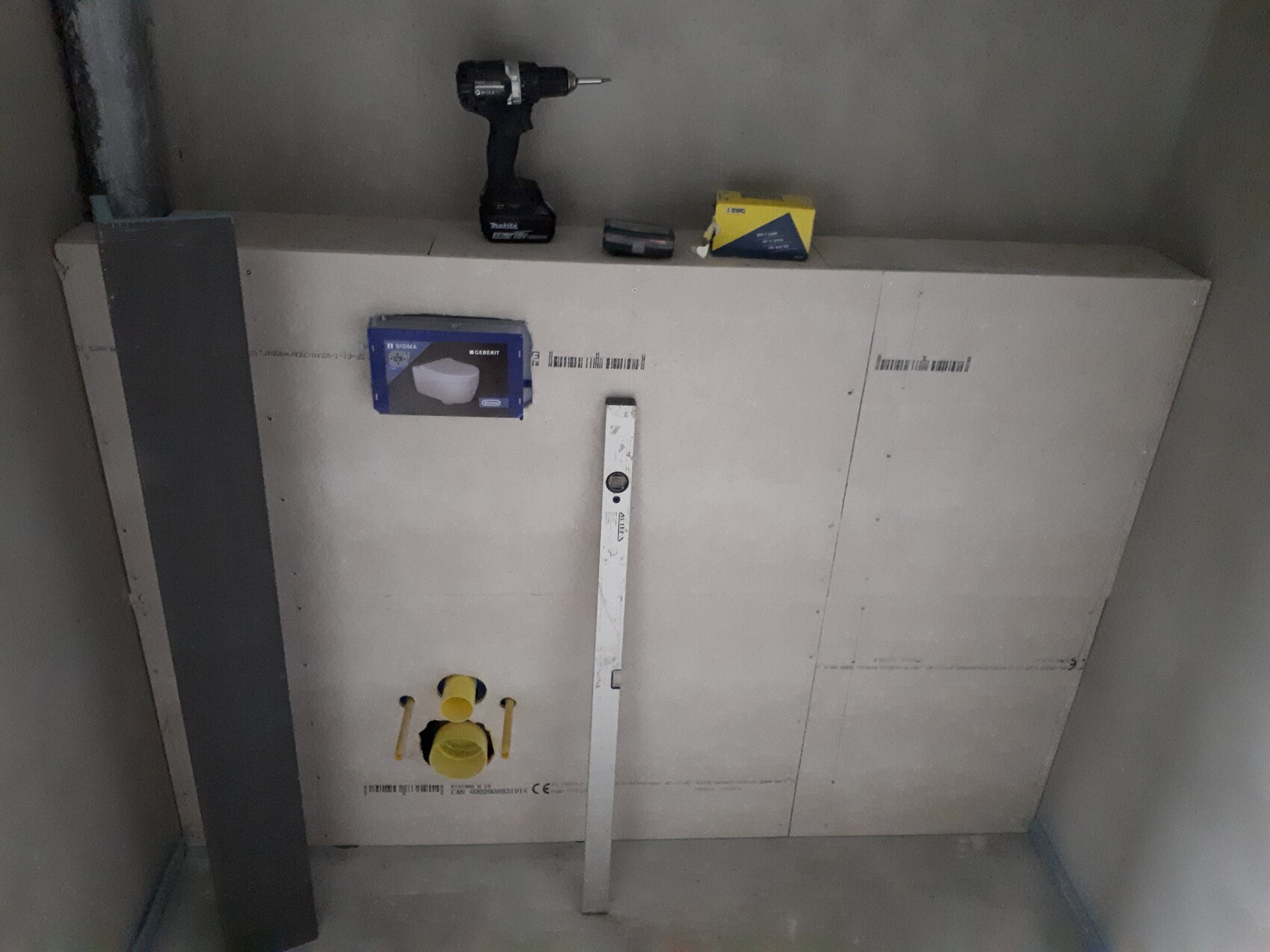 Bauwand mit Installationspanel, Bohrmaschine, Werkzeugbox, gelbem Hahnventil und Wasserwaage.
