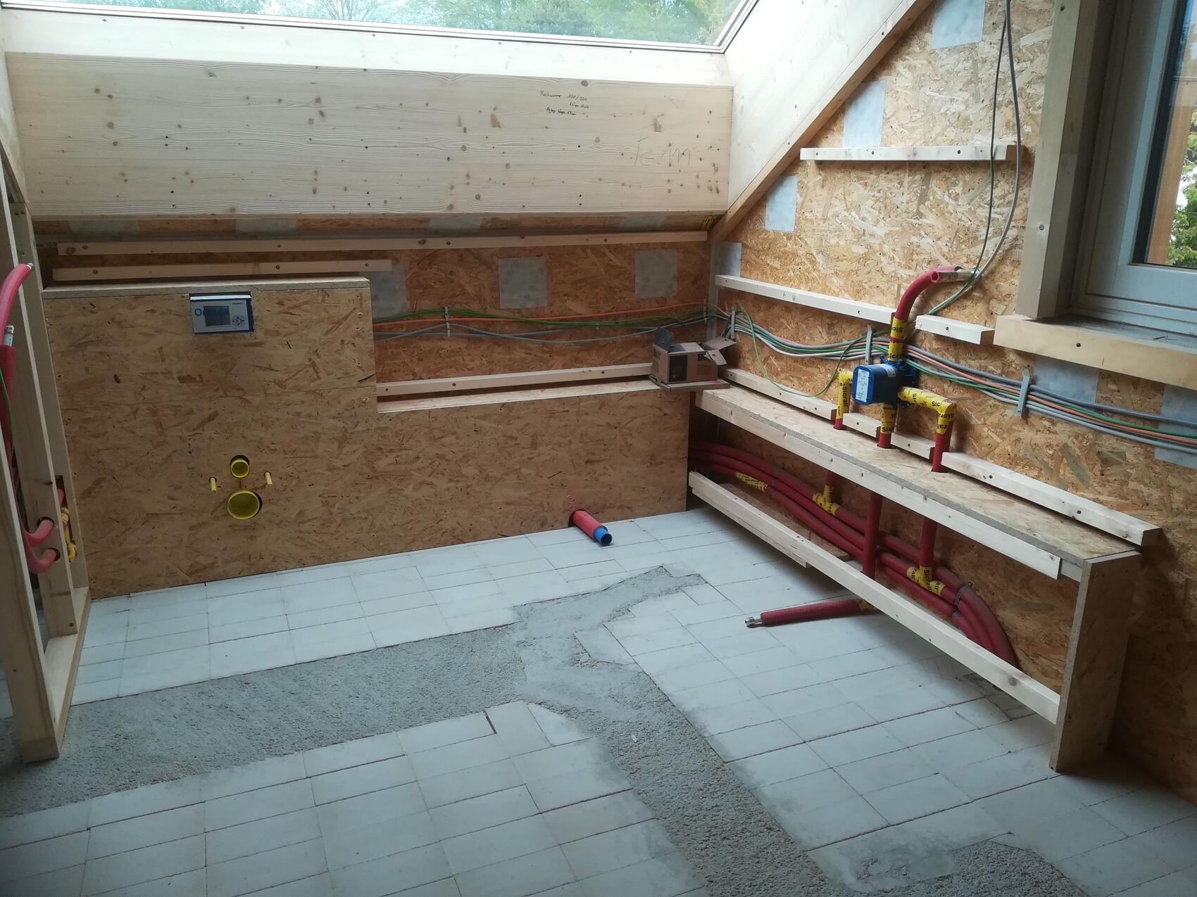 Rohbau-Badezimmer: OSB-Wände, rote Wasserleitungen, Holzregale, Fenster, Fliesenboden in Arbeit.