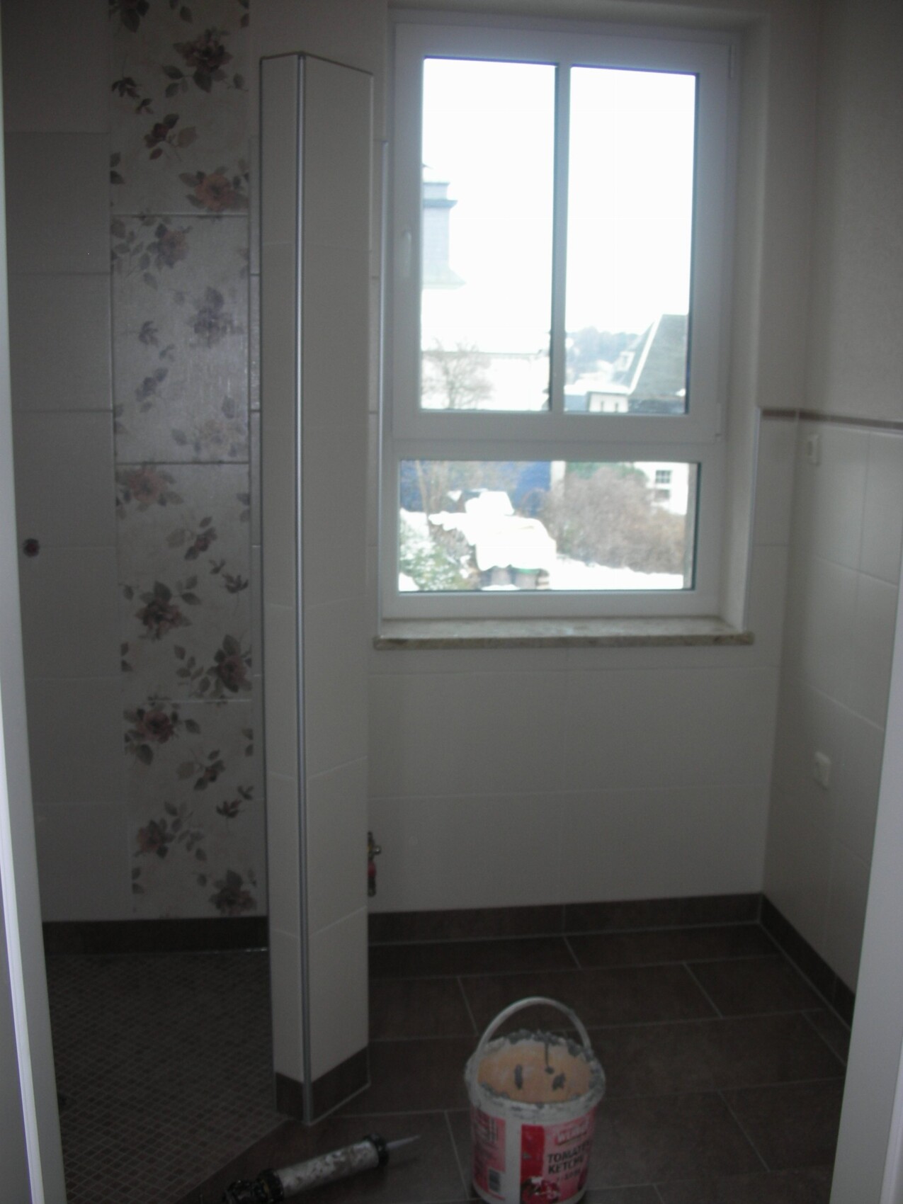 Badezimmer im Bauzustand mit Dusche, Fliesen, Fenster und Eimer auf dem Boden.