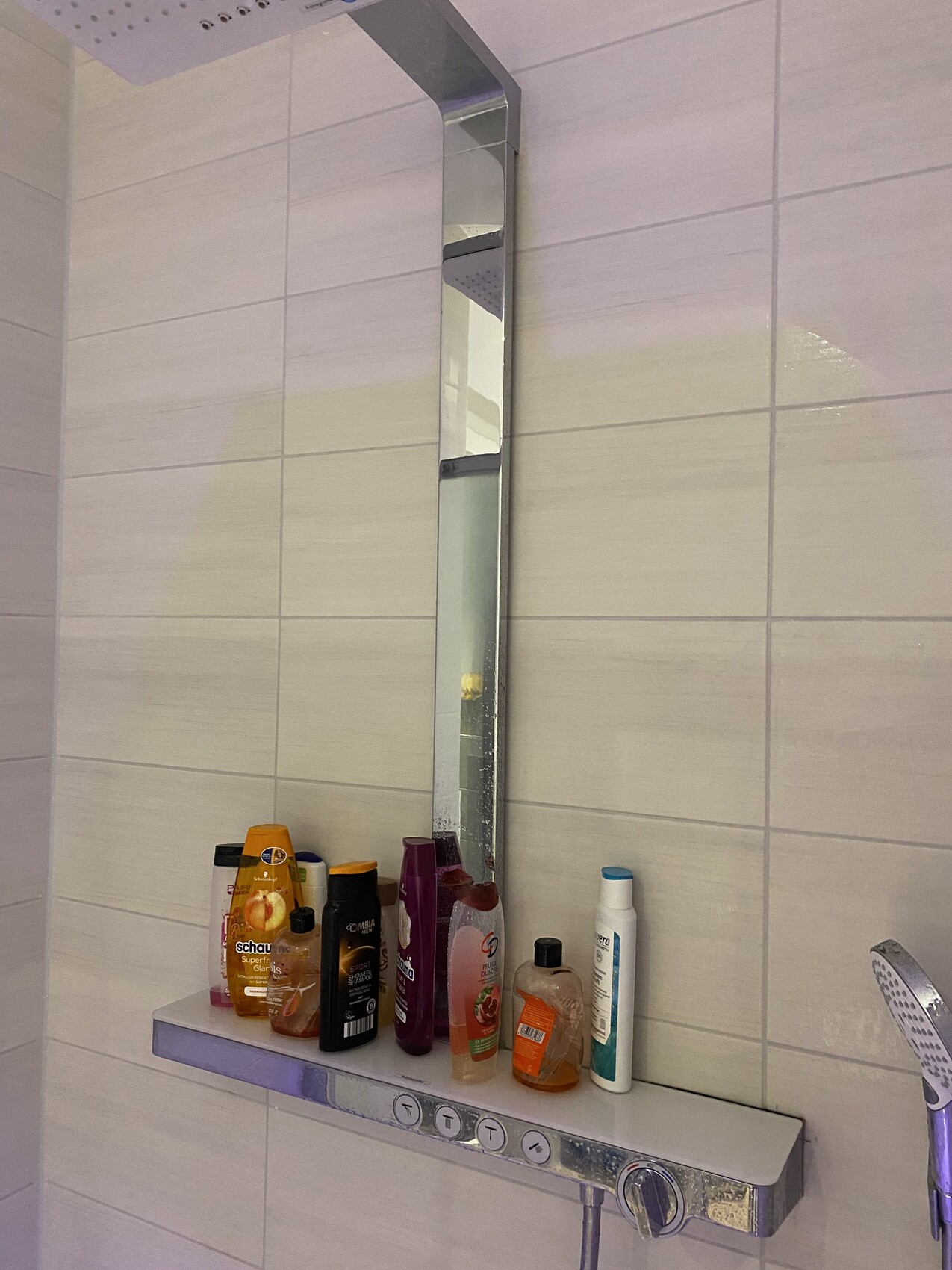 Duschbereich mit schmalem Spiegel und Ablage voller Shampoo, Duschgel, Lotion.