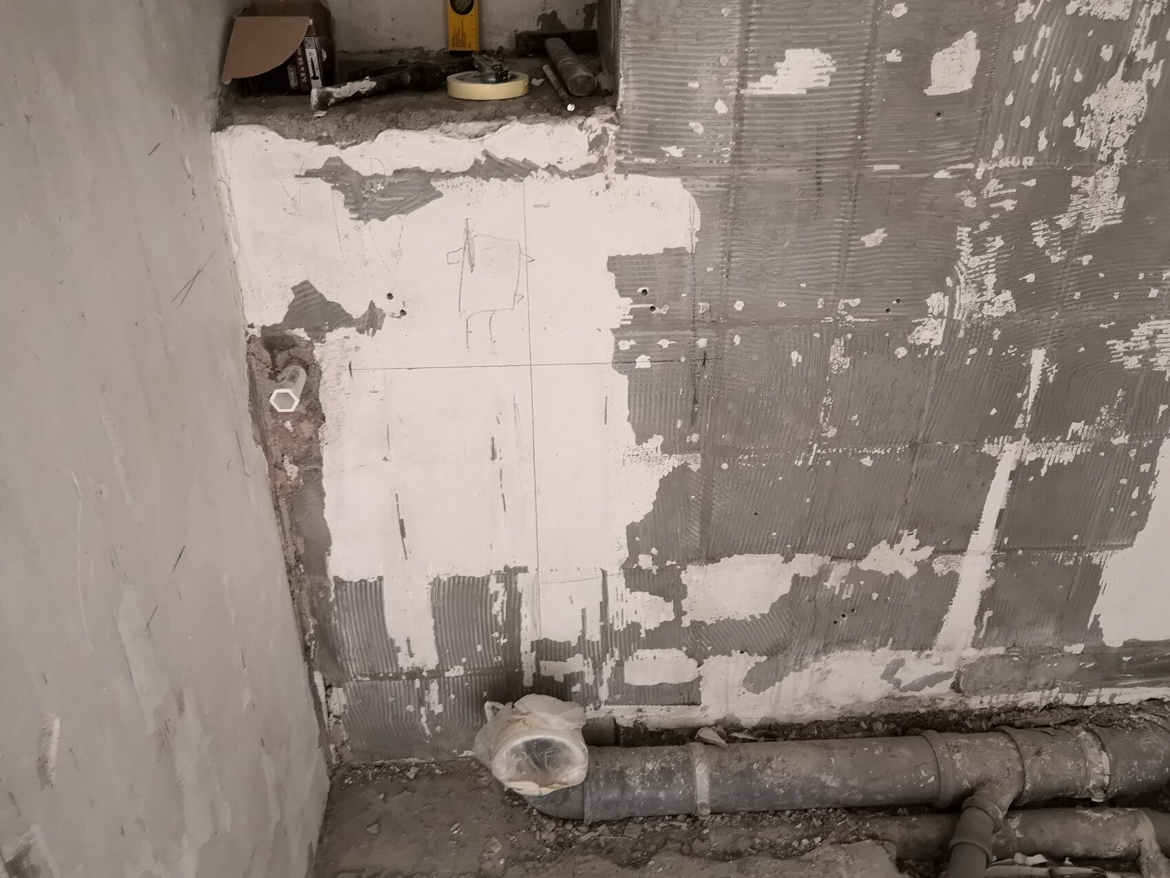 Baustelle: Wand mit teilweise abgelösten Fliesen, sichtbare Rohrleitungen am Boden.