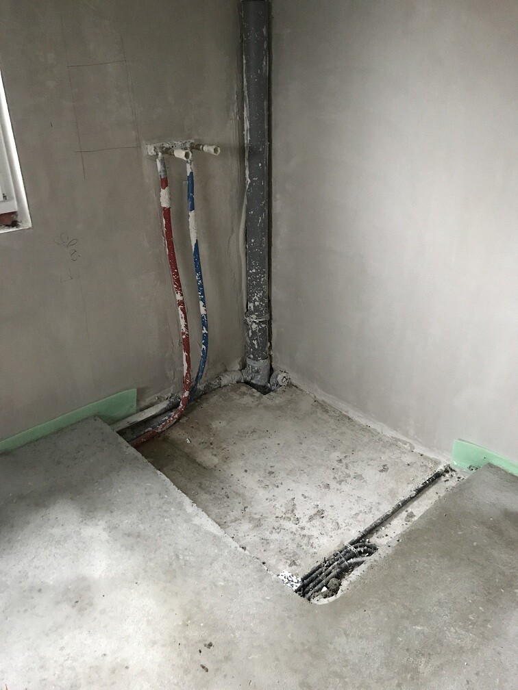 Baustellen-Ecke: rote und blaue Wasserleitungen, senkrechtes Rohr, offener Bodenkanal mit Gitter.
