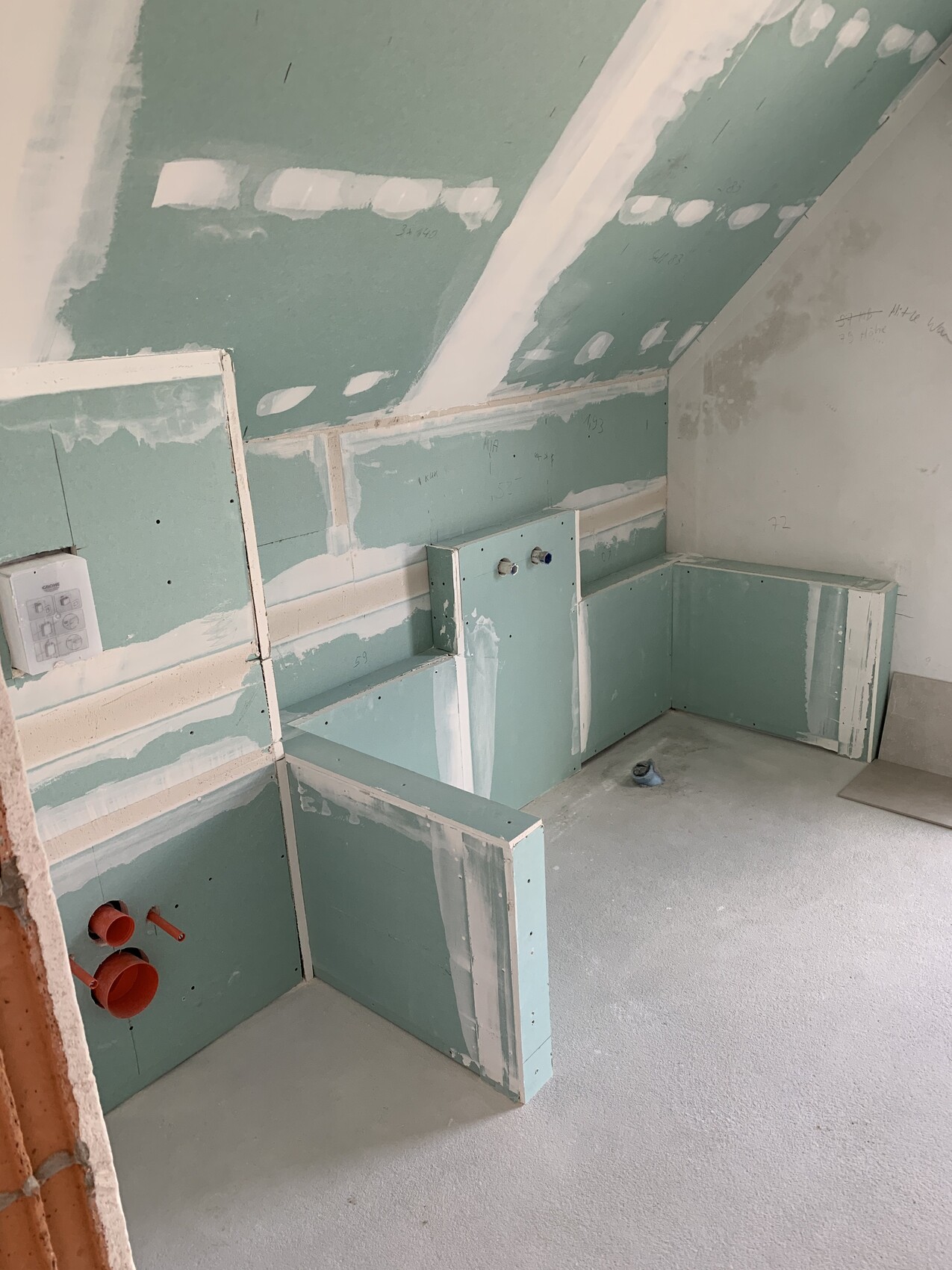 Unfertiger Innenraum mit grünen Gipskartonplatten, schräger Decke und Sanitärrohren.