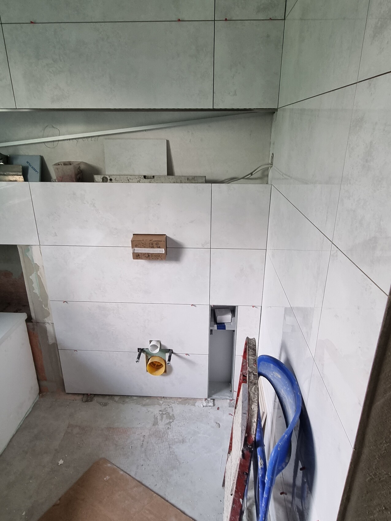 Badezimmer im Rohbau: Fliesen, gelber Abfluss, Sanitäranschlüsse, blaue Schlauchleitung.
