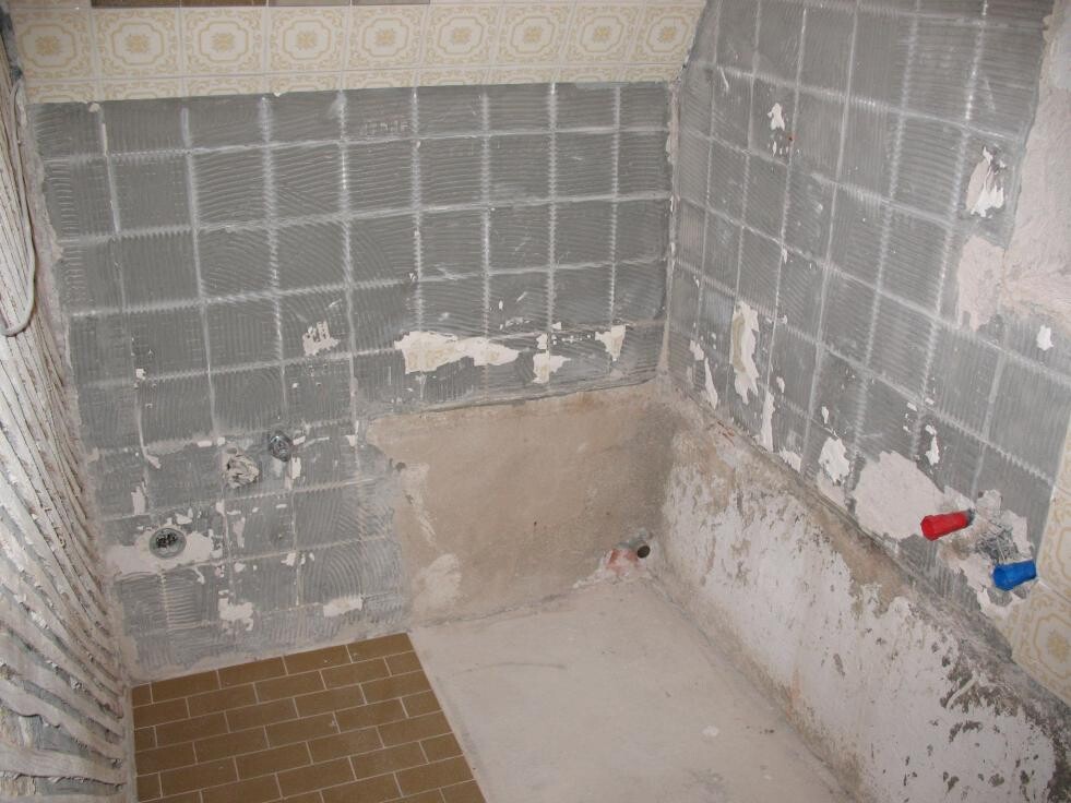 Badezimmer im Renovierungszustand mit grauen Fliesen, offenen Rohren und Bauarbeiten.