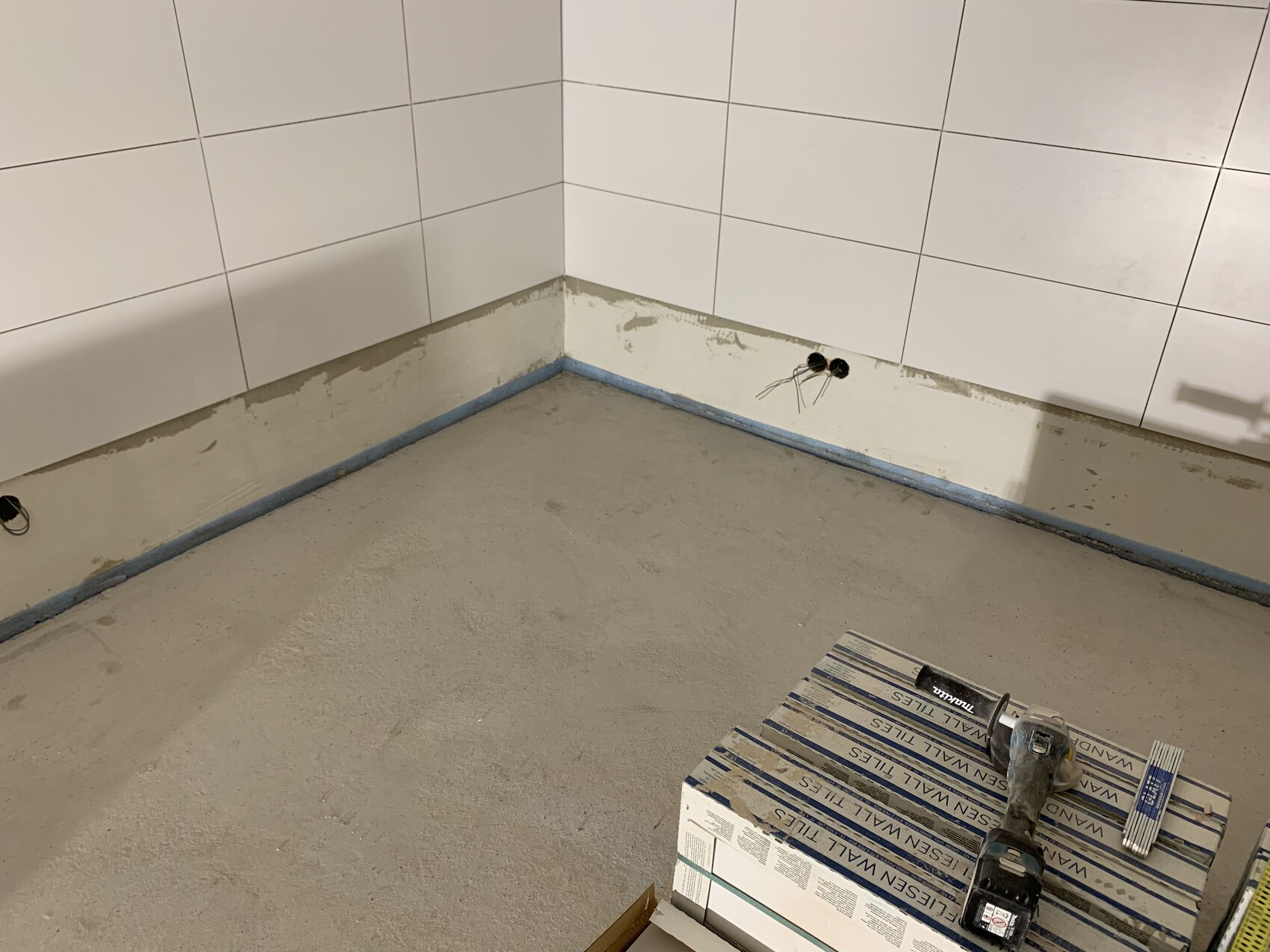 Ecke eines renovierten Raums: weiße Fliesen, freiliegende Leitungen, Bohrmaschine auf Kisten.