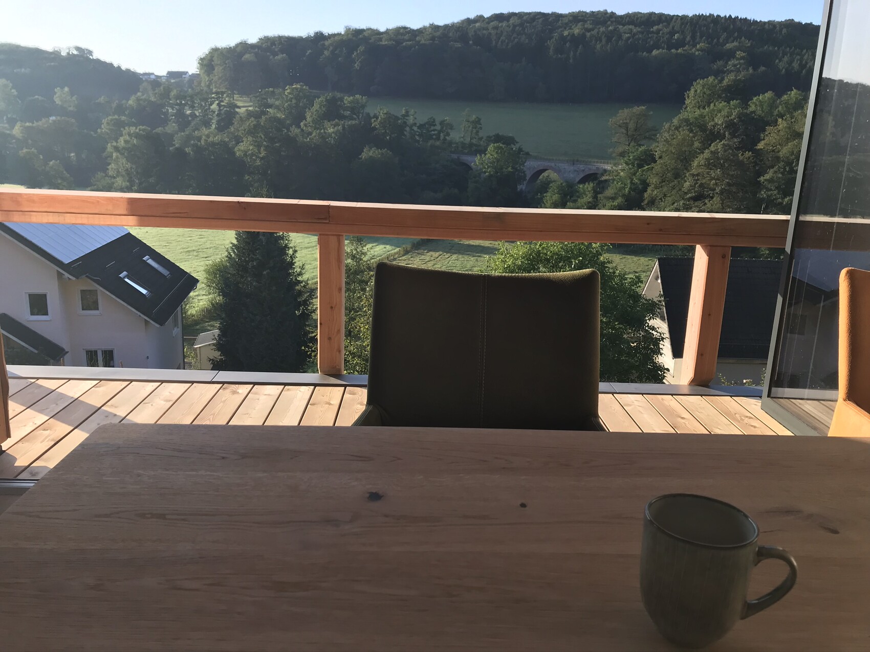 Holzterrasse mit Tisch und Kaffeetasse im Vordergrund, Blick auf grüne Landschaft und Brücke im Tal.