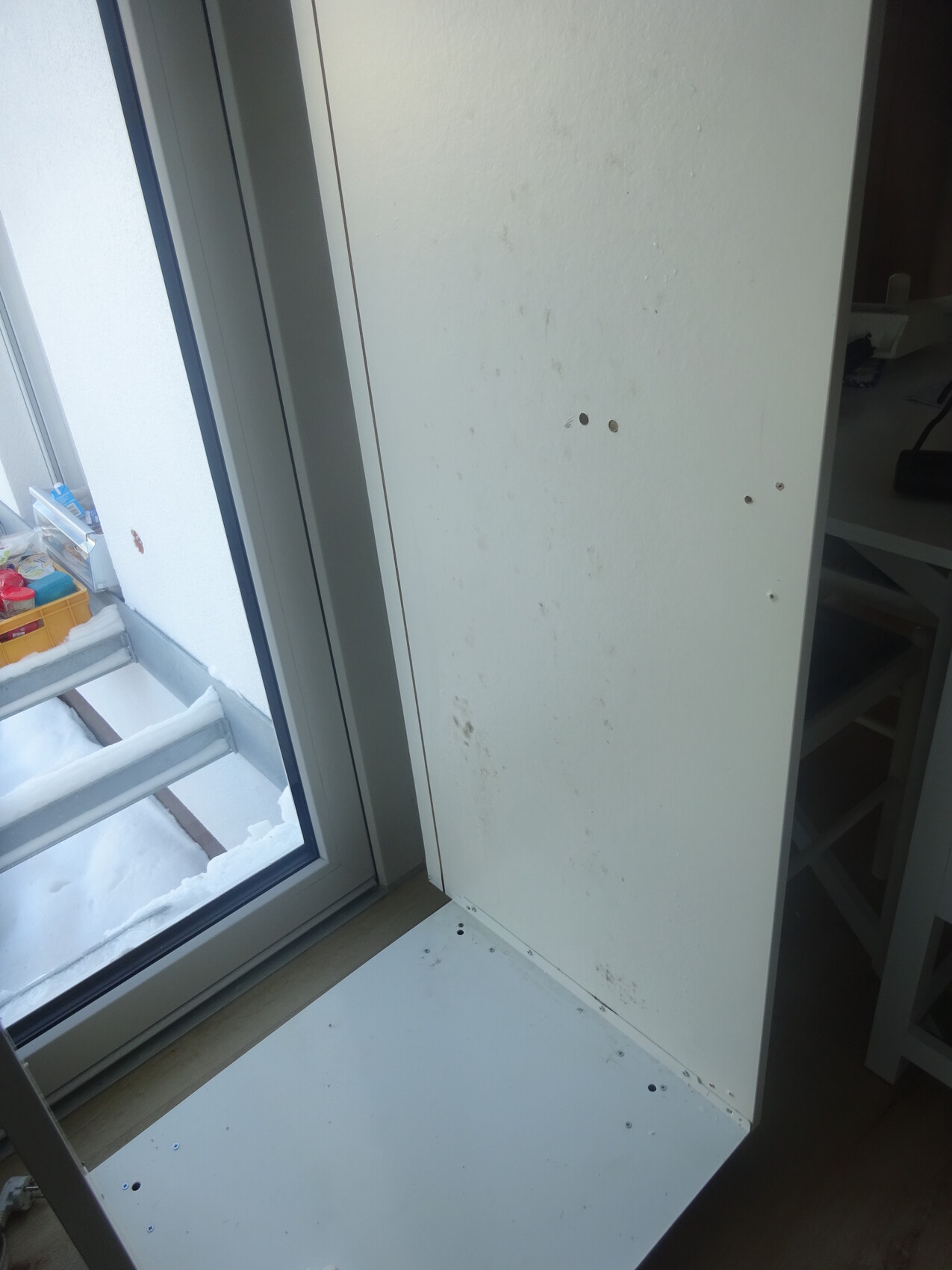 Weißes Möbelpaneel mit Flecken und Bohrlöchern neben einem Fenster.