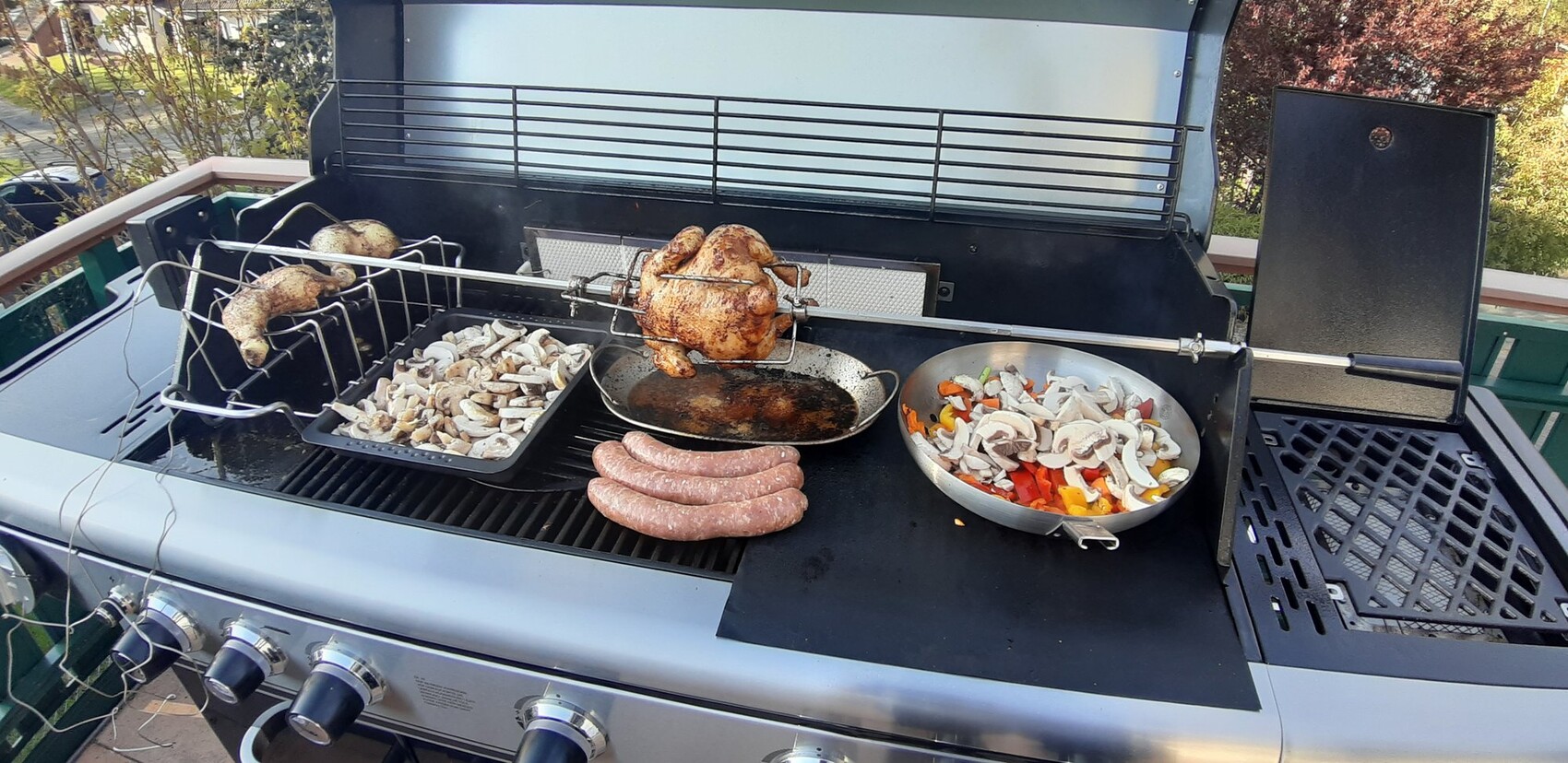 Grill auf Balkon: Hähnchen am Drehspieß, Würstchen, Pilze und buntes Gemüse auf der Grillfläche.