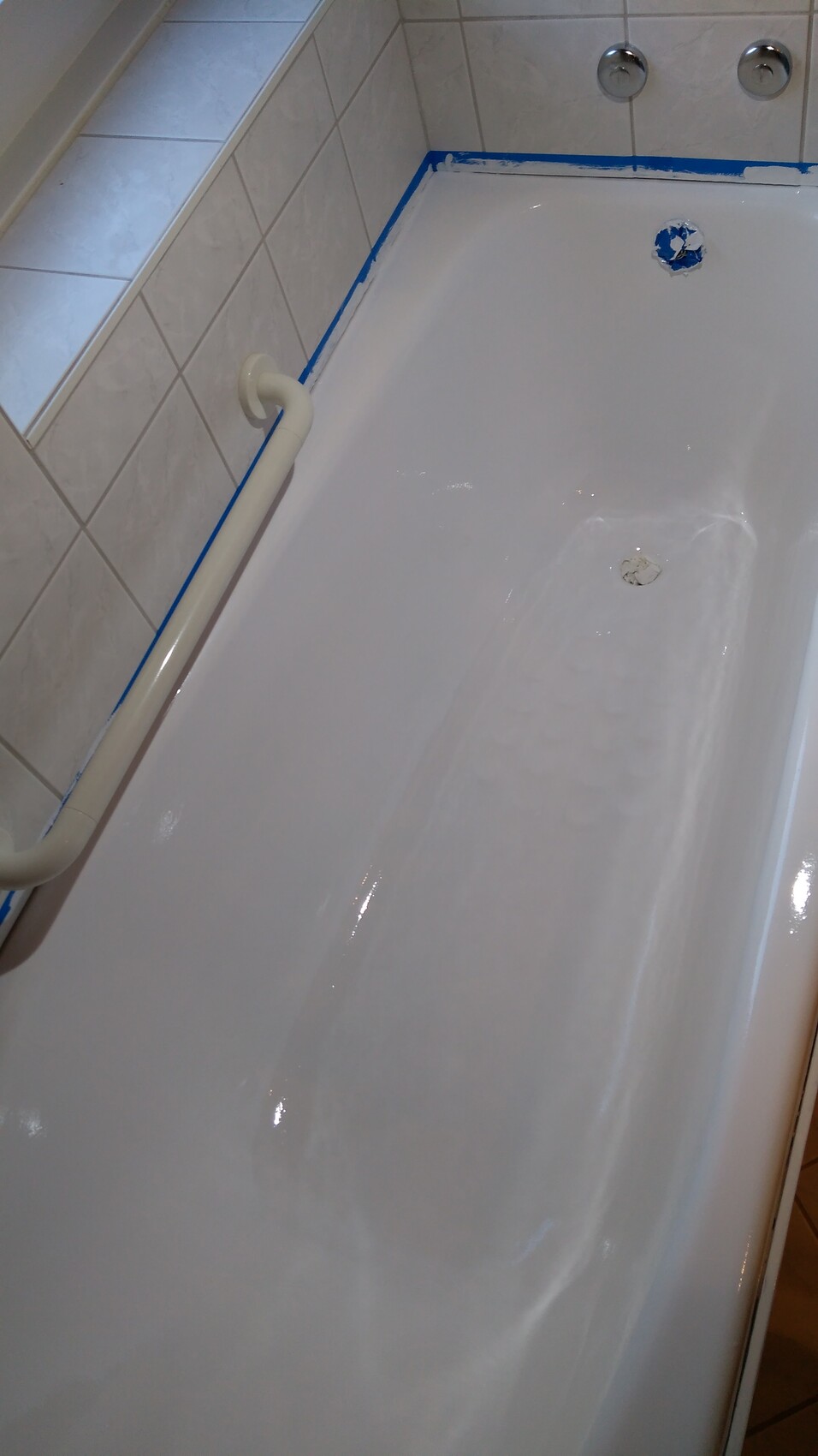 Badezimmer-Badewanne mit Haltegriff, Fliesenwand und blauem Malerband am Rand.