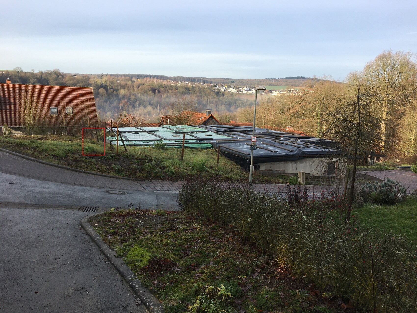 Straße im Vorort mit roten Ziegeldächern; Baufläche mit Plane im Zentrum und Hügel im Hintergrund.