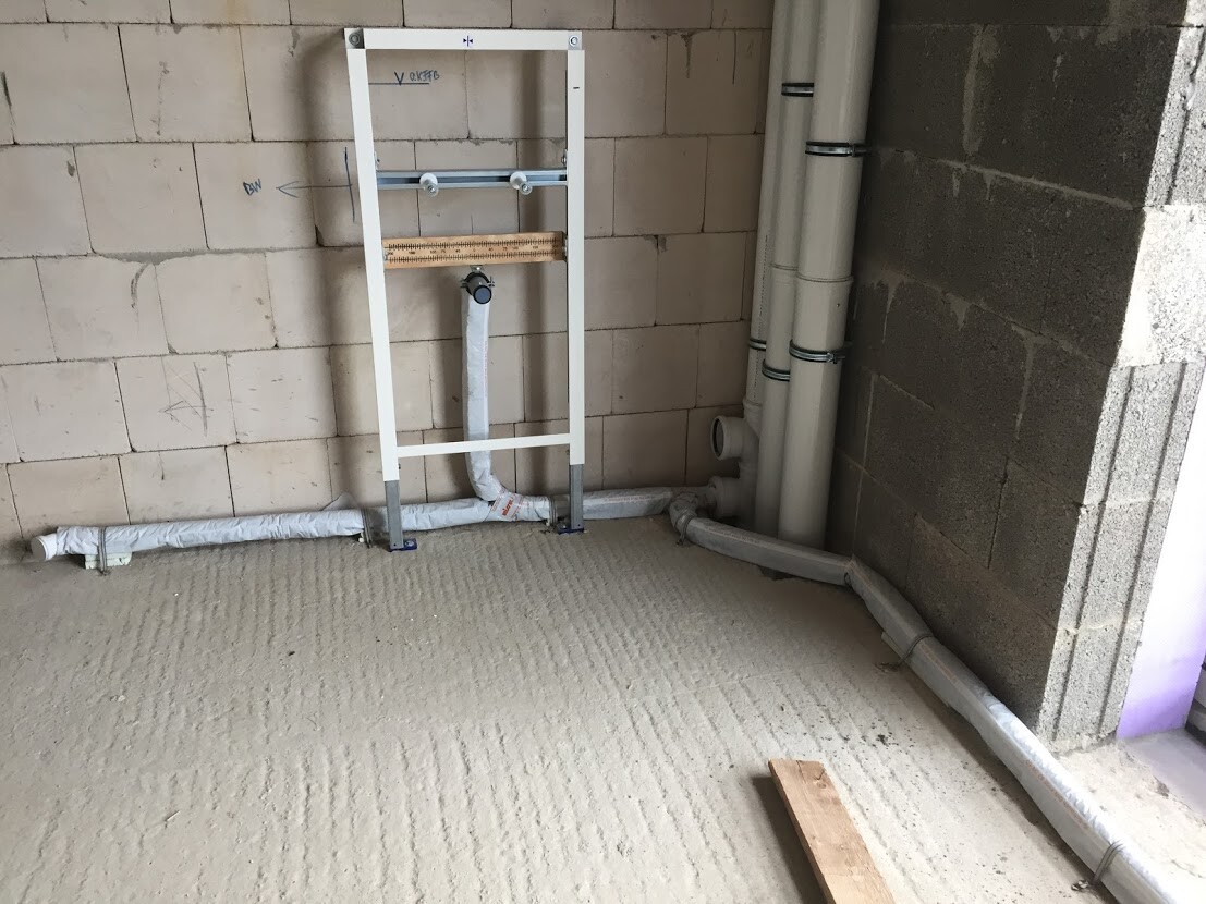 Rohbau-Bad: Weißer WC-Rahmen, Abflussrohre und Wasserleitungen an Wand und Boden.