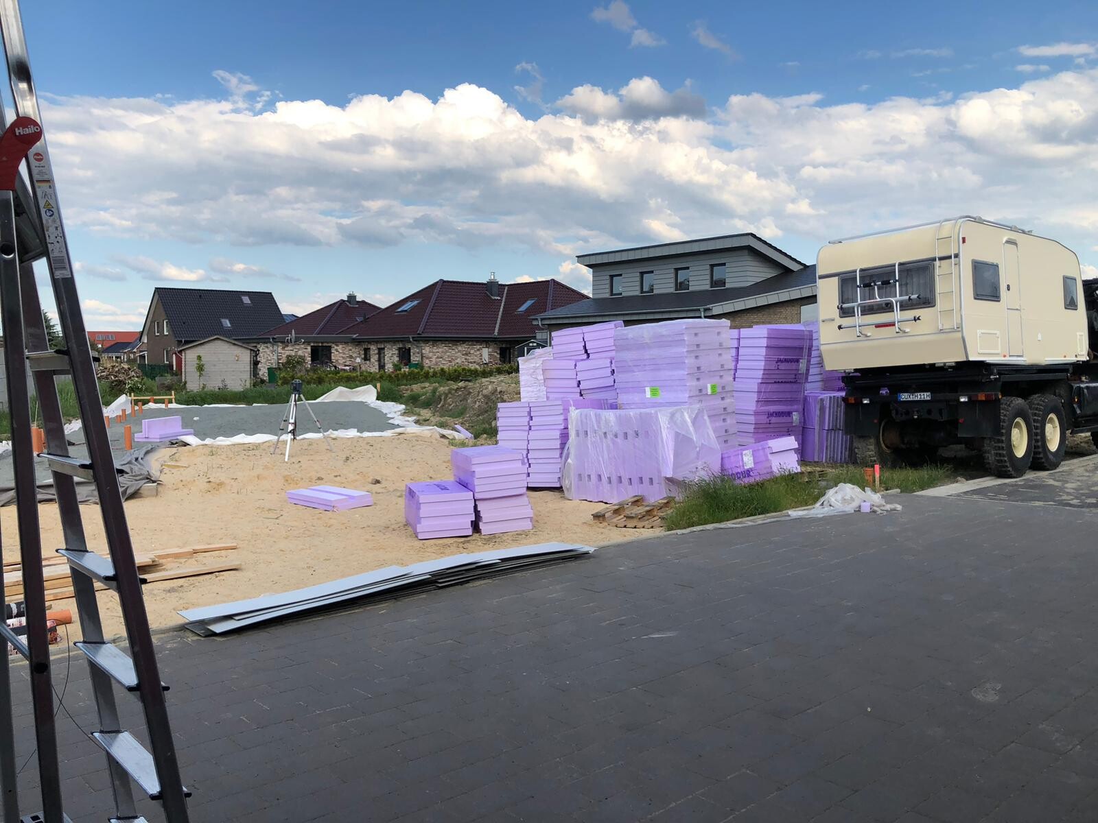 Baustelle mit gestapelten violetten Dämmplatten, Bau-LKW und Häusern im Hintergrund.