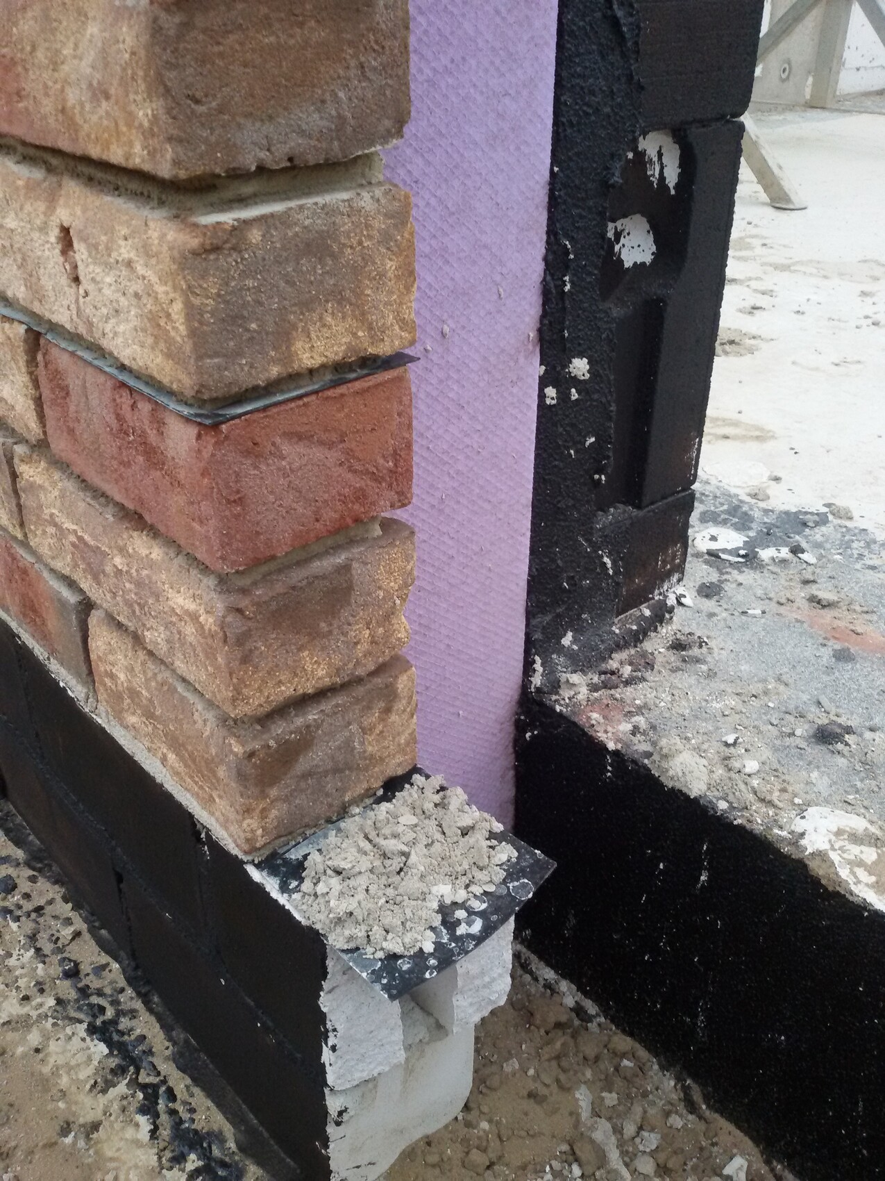 Baustelle: Ziegelwand mit rosa Dämmplatte, schwarze Abdichtung, Geröll am Fundament.