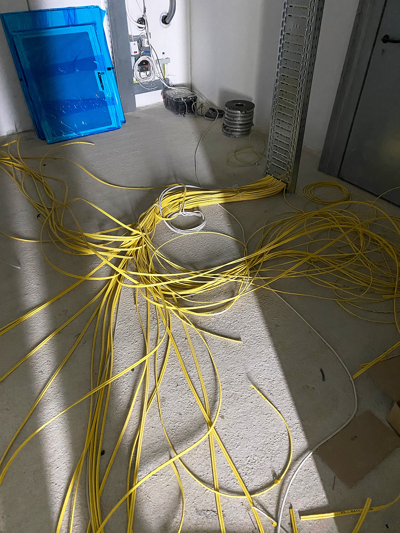 Gelbe Kabel liegen kreuz und quer auf Betonboden in einem Technikraum; links eine blaue Kiste.