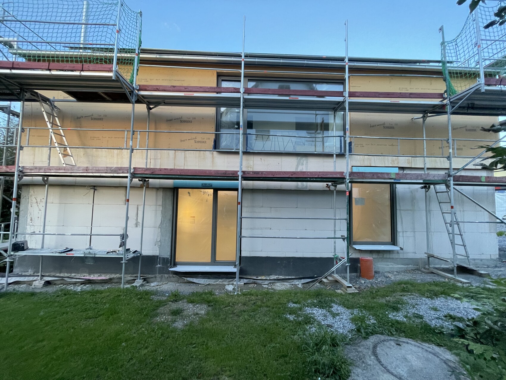 Mehrstöckiges Haus unter Bau mit Gerüst um die Fassade, Fensterfronten und Grünfläche davor.
