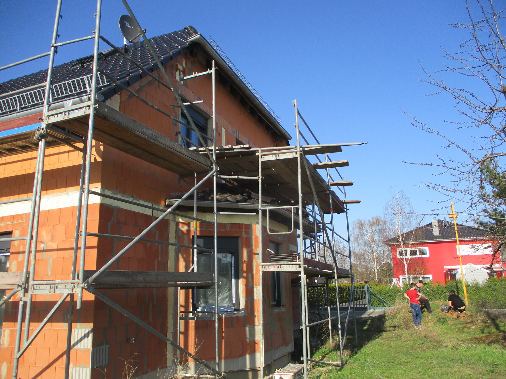 Rohbau eines orange Ziegelhauses mit Gerüst; Bauarbeiter im Garten, blauer Himmel.
