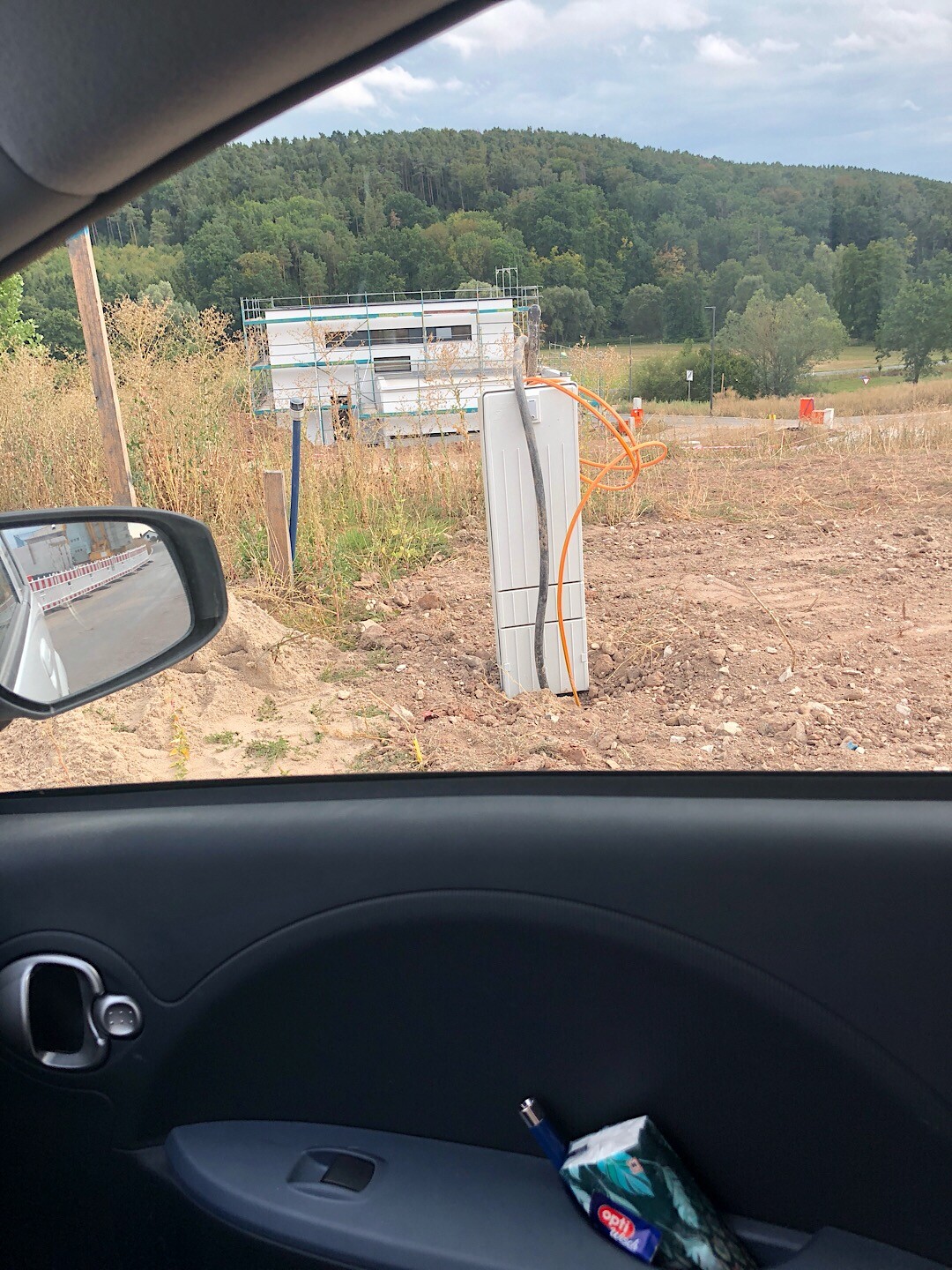 Baustelle mit freier Elektro-Verteilerbox, orange Kabel, Auto-Fenster im Vordergrund, Wald im Hintergrund