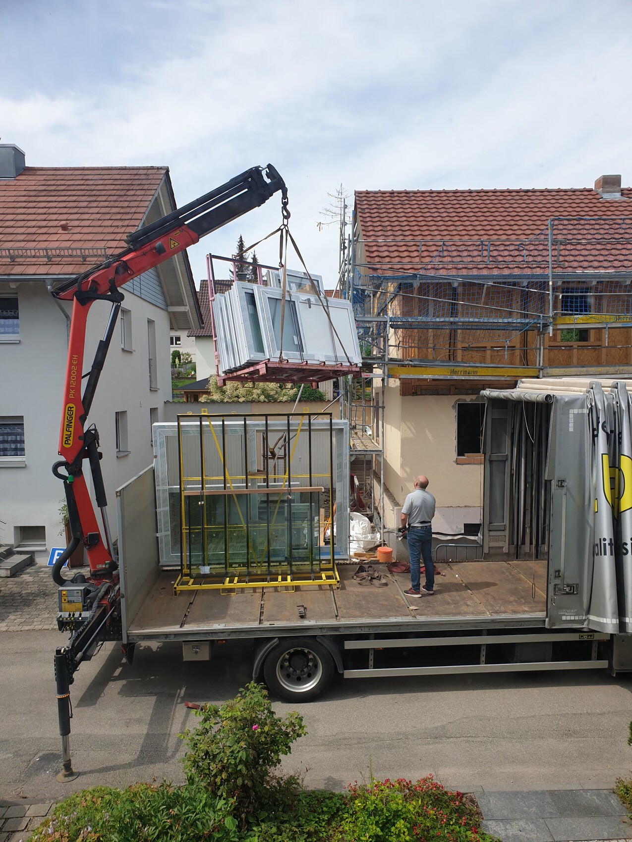 Kran hebt Fensterelemente auf Baugerüst eines Wohnhauses; Arbeiter auf LKW-Plattform.