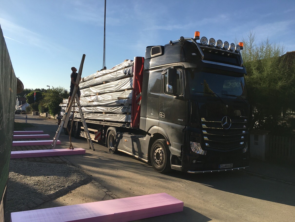 Großer Lastwagen belädt Baulieferung mit langen Holzbalken auf einer Baustelle.