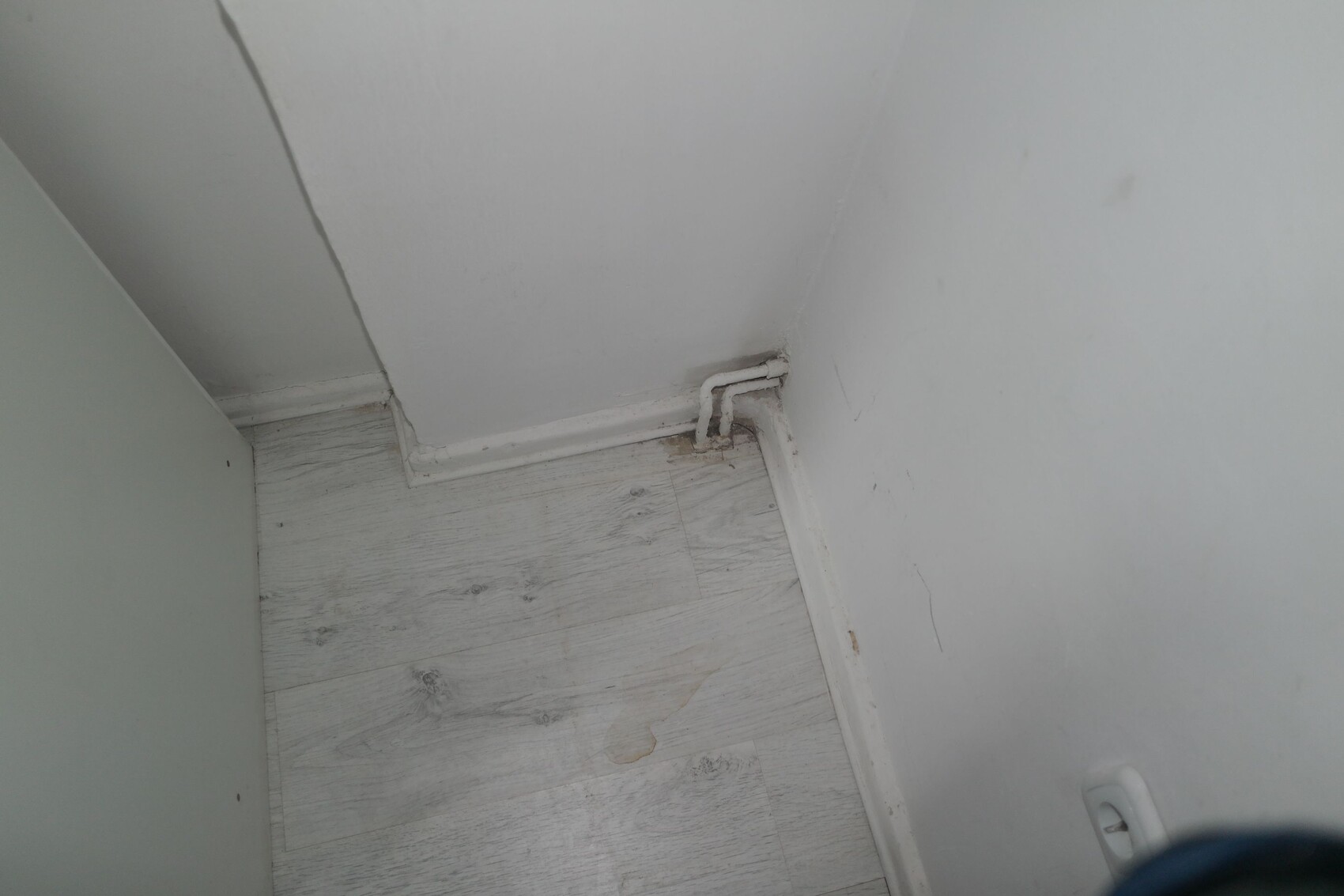 Eckansicht eines Zimmers: weiße Wände, grauer Laminatboden, eine kurze Rohrleitung am Boden.