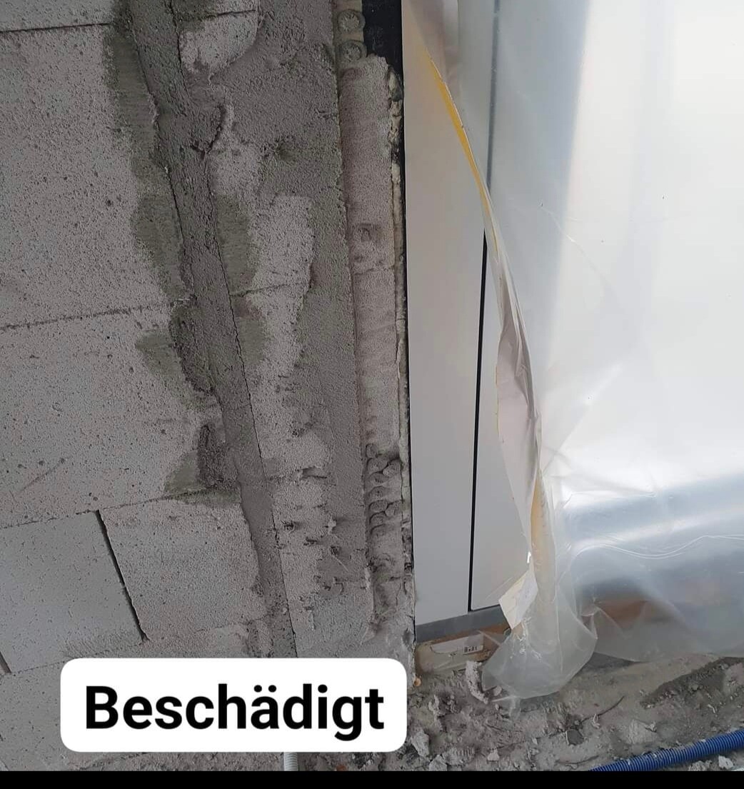 Beschädigte Betonwand mit freiliegendem Mauerwerk, Bauabfall und Plastikfolie an einer Tür.