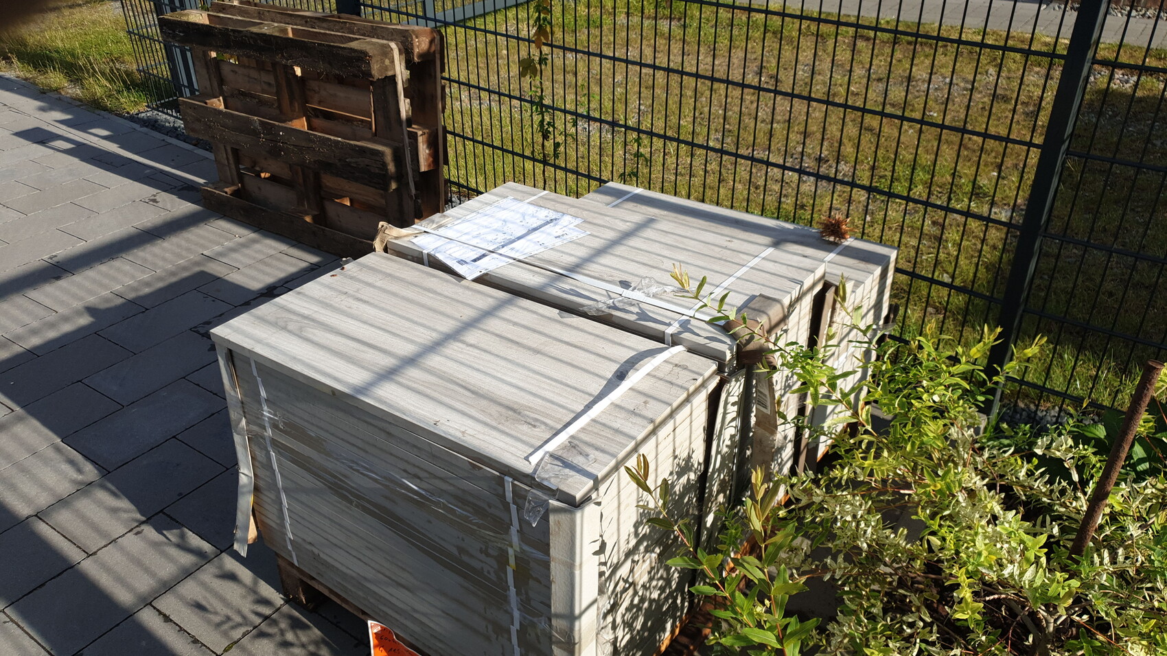 Große Paletten mit verpackten Bauteilen stehen auf dem Gehweg neben einem Metallzaun im Freien.