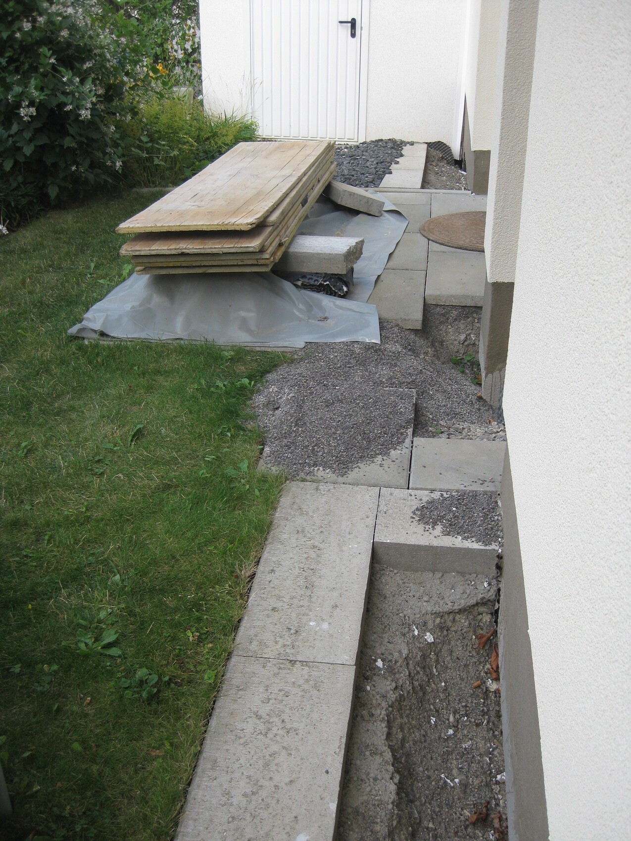 Baustellenmaterialien wie Holzplatten, Pflastersteine und Kies liegen auf einer Plane vor einem Haus.