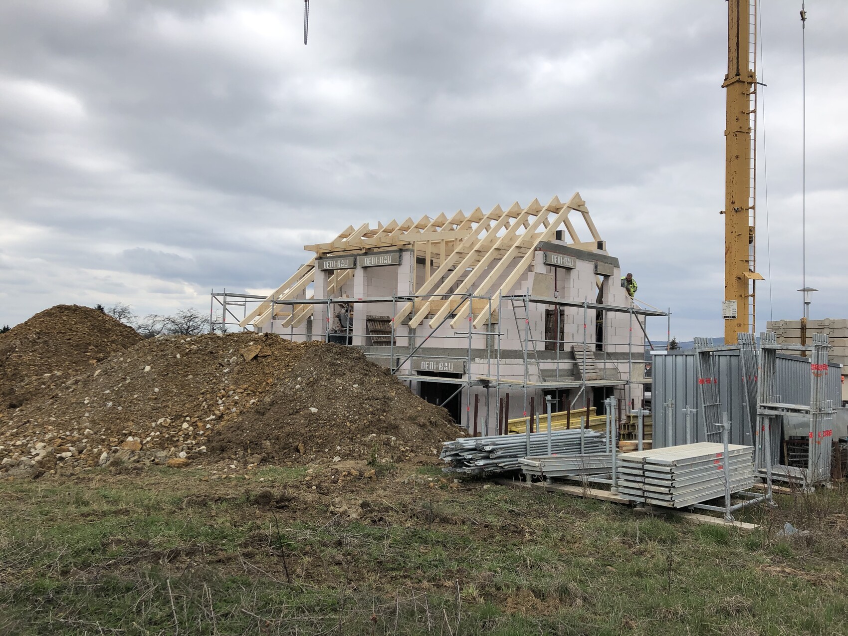 Baustelle mit halbfertigem Holzhaus, Dachstuhl, Gerüst, Kran und Materialstapeln.