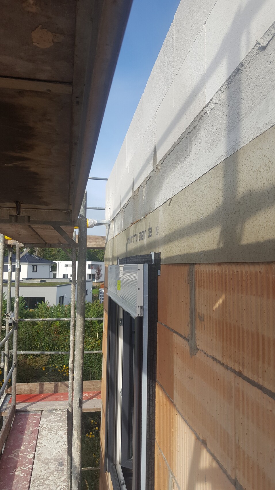 Baugerüst am Neubau; Fensterrahmen neben einer Ziegelwand, Blick auf Nachbarhäuser.