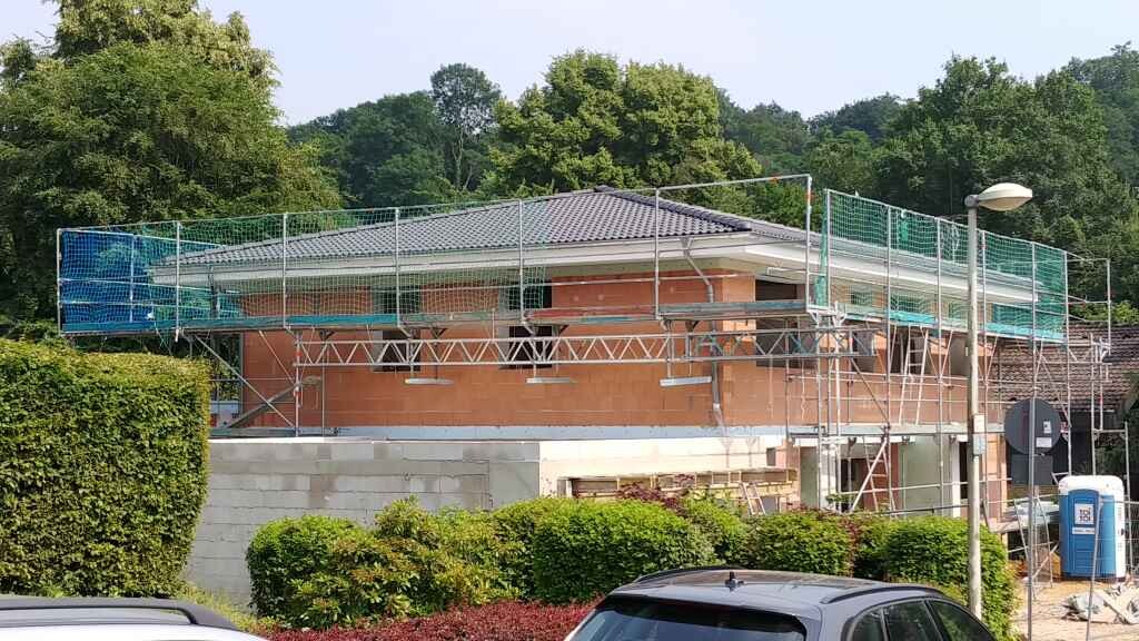 Ein Haus mit Gerüst, orange Backsteinfront, grüne Netze; Baustelle und Bäume dahinter.