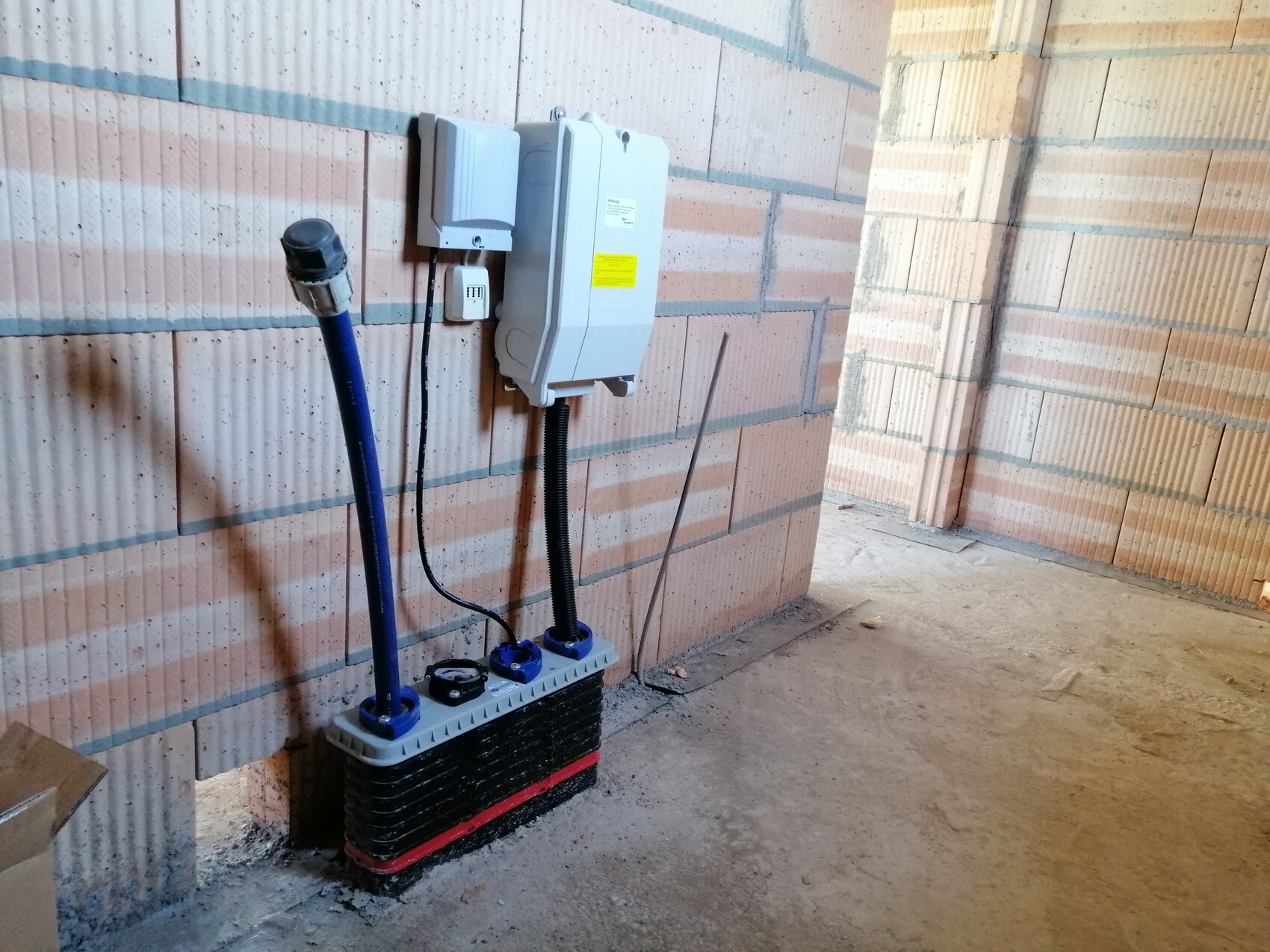 Baustelle: Wandmontiertes Elektrogerät mit Warnetikett, blaue Schläuche führen zu schwarzem Kasten.