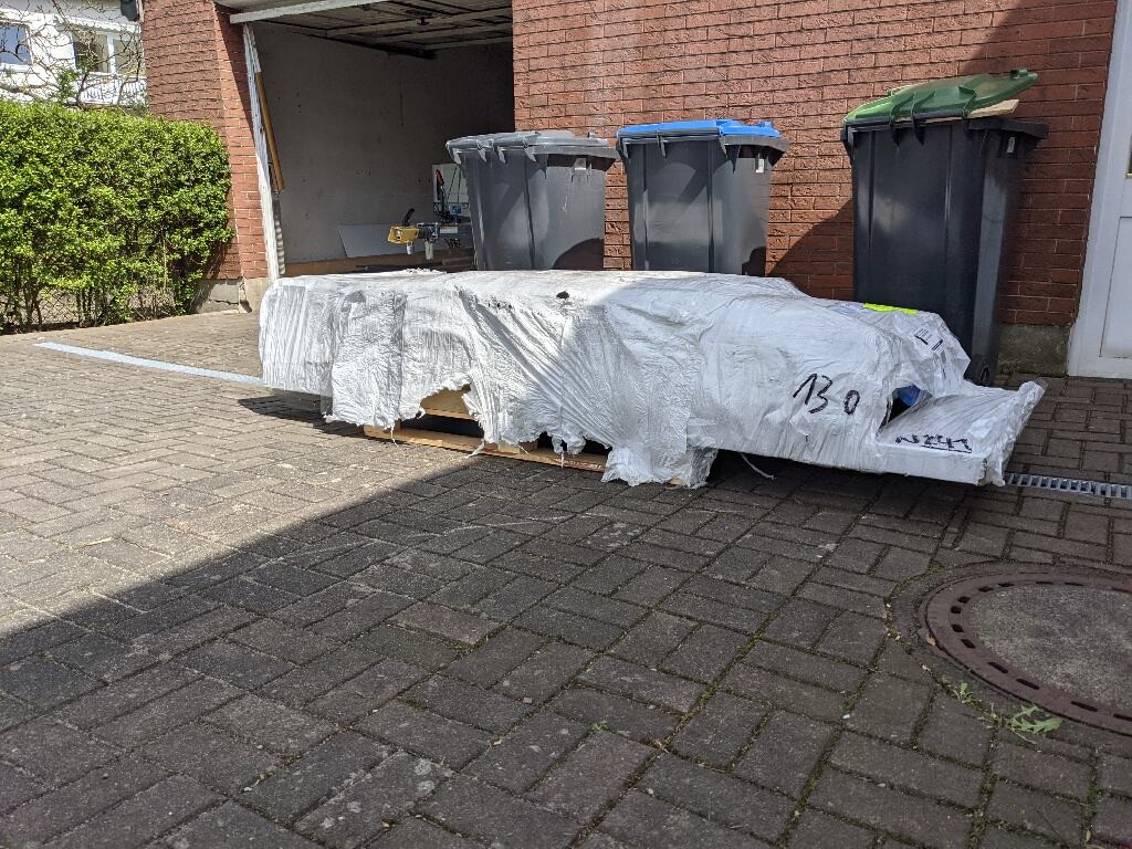 Lange, mit Plane bedeckte Palette auf dem Hof vor der Garage; Mülltonnen dahinter.