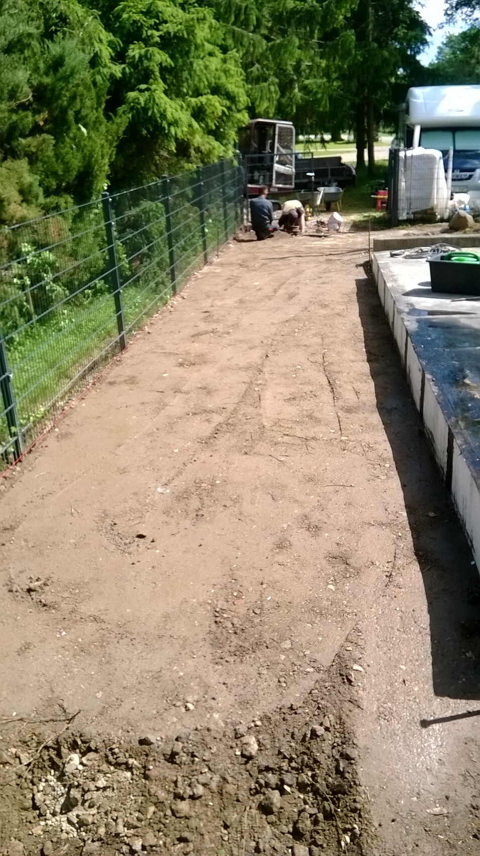 Zwei Arbeiter arbeiten am Boden neben einem Metallzaun; im Hintergrund ein Lastwagen.