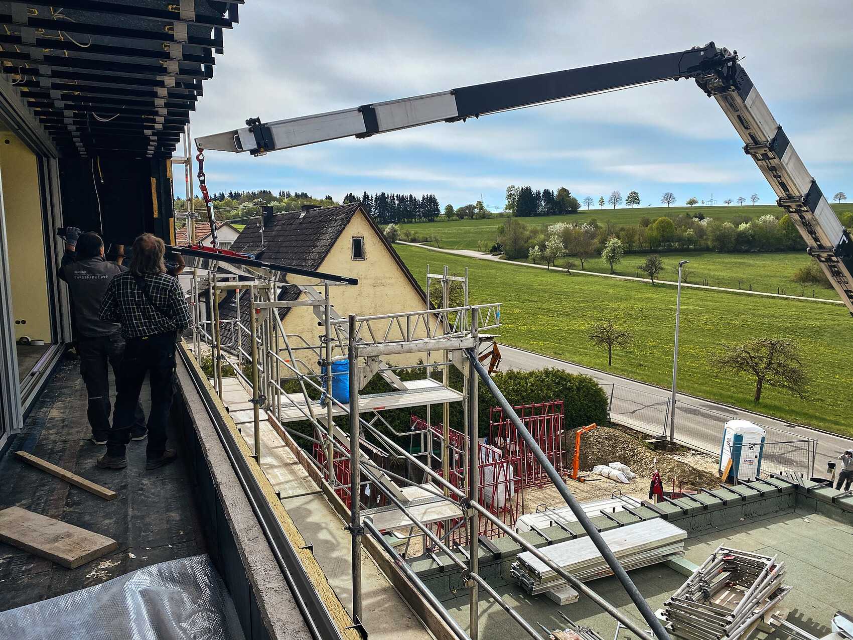 Baustelle: Gerüst, Kranarm, zwei Arbeiter an einer Bauwand, Blick auf grüne Landschaft.